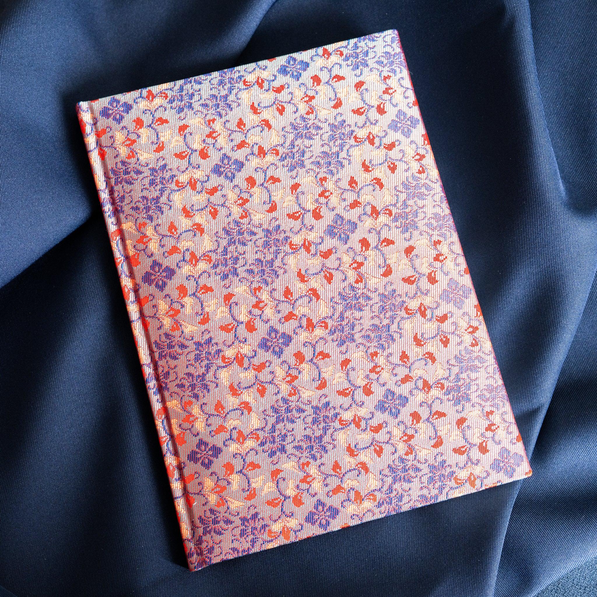Yusoku Hanabishi Nishijin silk notebook angled view, full blue and red woven floral cover, no belly band, on blue fabric