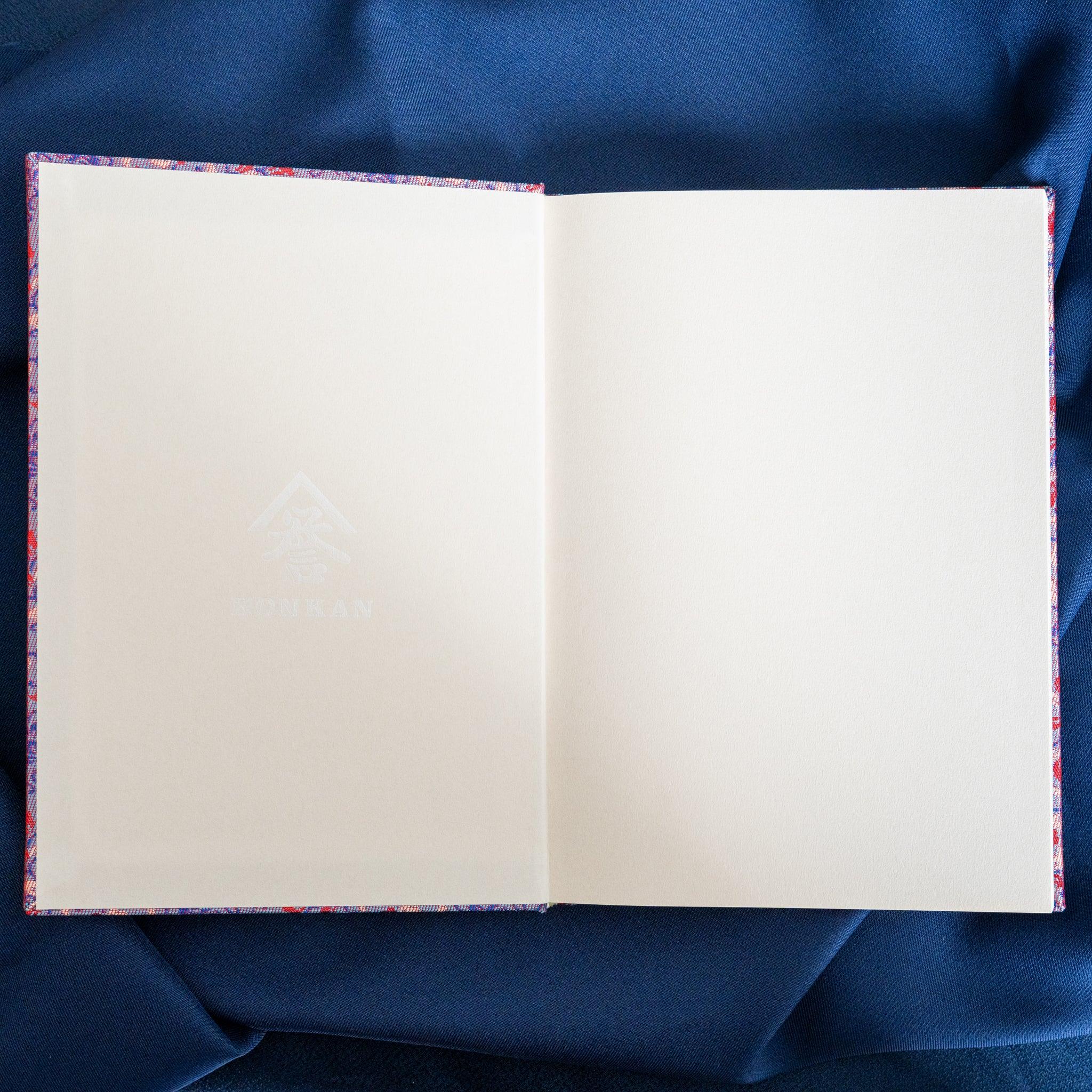 Yusoku Hanabishi Nishijin silk notebook open flat, two blank white pages spread, silk cover edges visible, on blue fabric