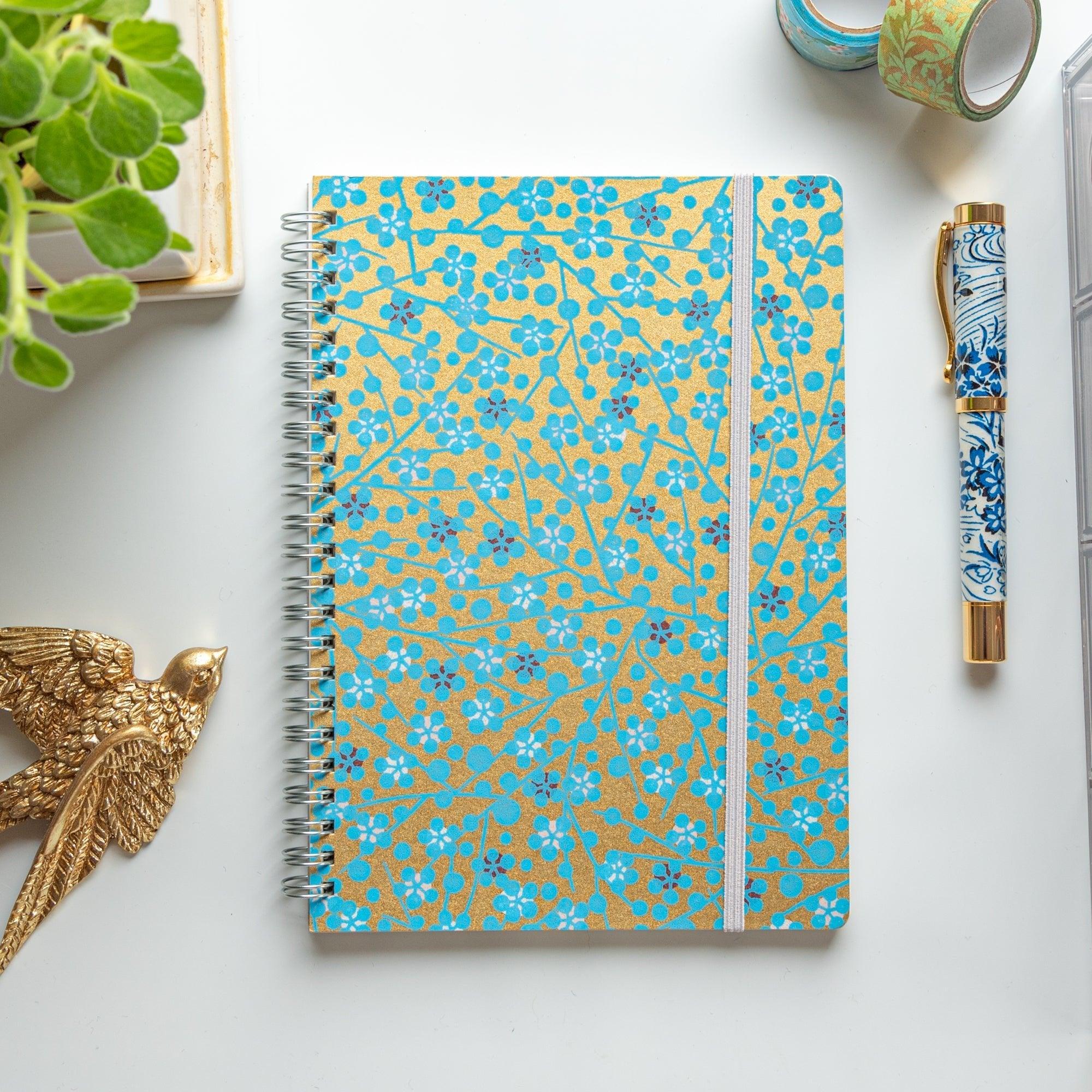 A Japanese notebook with a Yuzen washi cover featuring blue floral patterns on a gold background, displayed on a white desk.