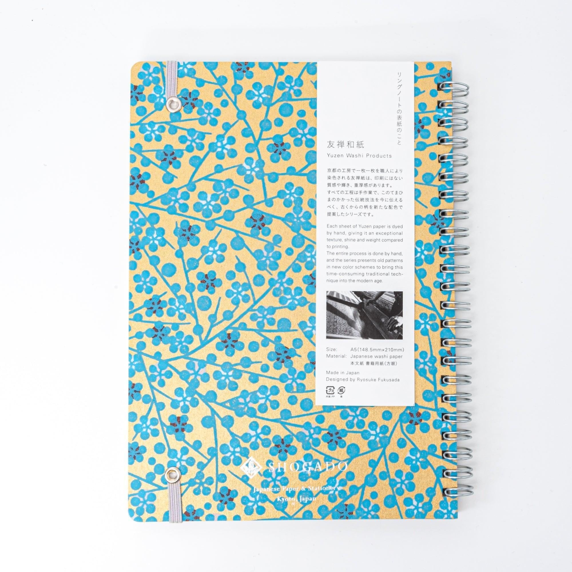 A Japanese notebook with a Yuzen washi cover featuring blue floral patterns on a gold background, displayed on a white desk.