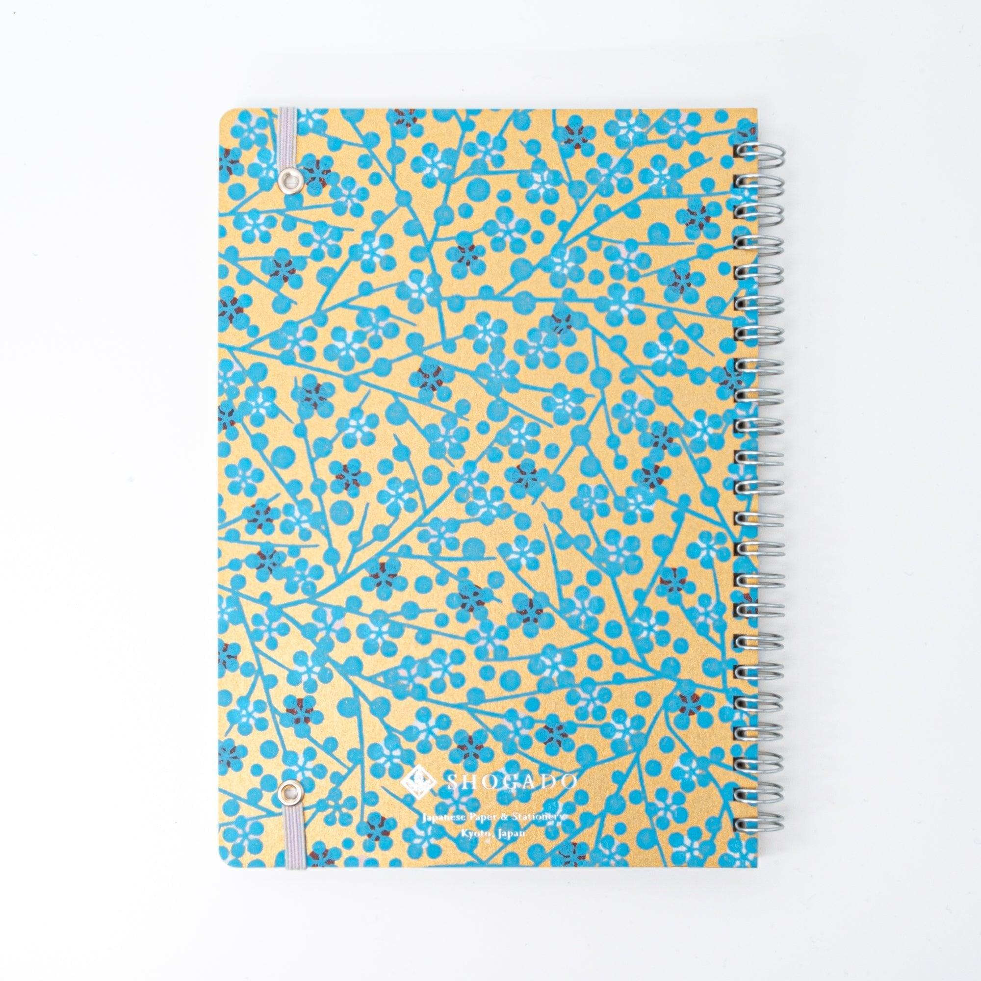 A Japanese notebook with a Yuzen washi cover featuring blue floral patterns on a gold background, displayed on a white desk.
