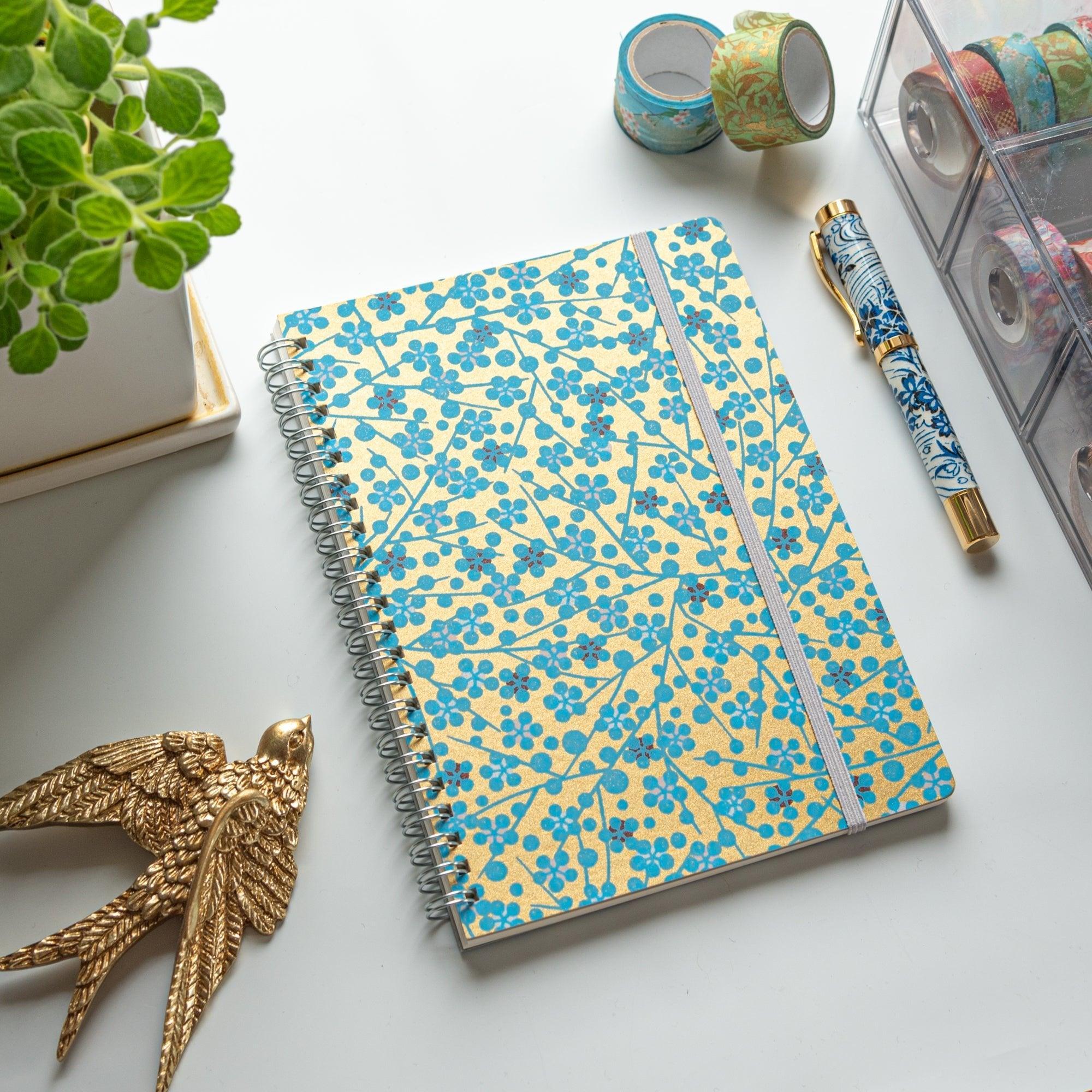 A Japanese notebook with a Yuzen washi cover featuring blue floral patterns on a gold background, displayed on a white desk.