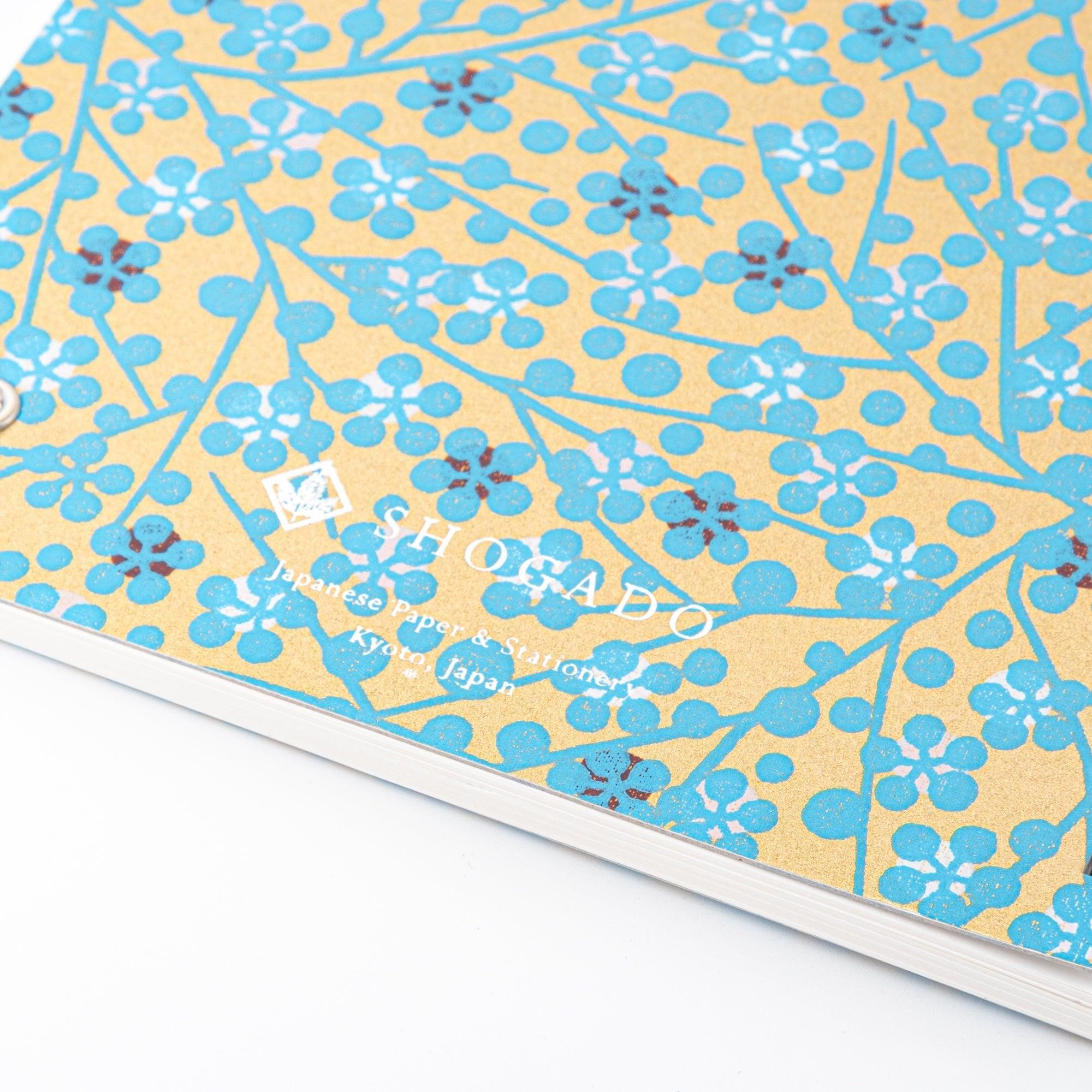 A Japanese notebook with a Yuzen washi cover featuring blue floral patterns on a gold background, displayed on a white desk.