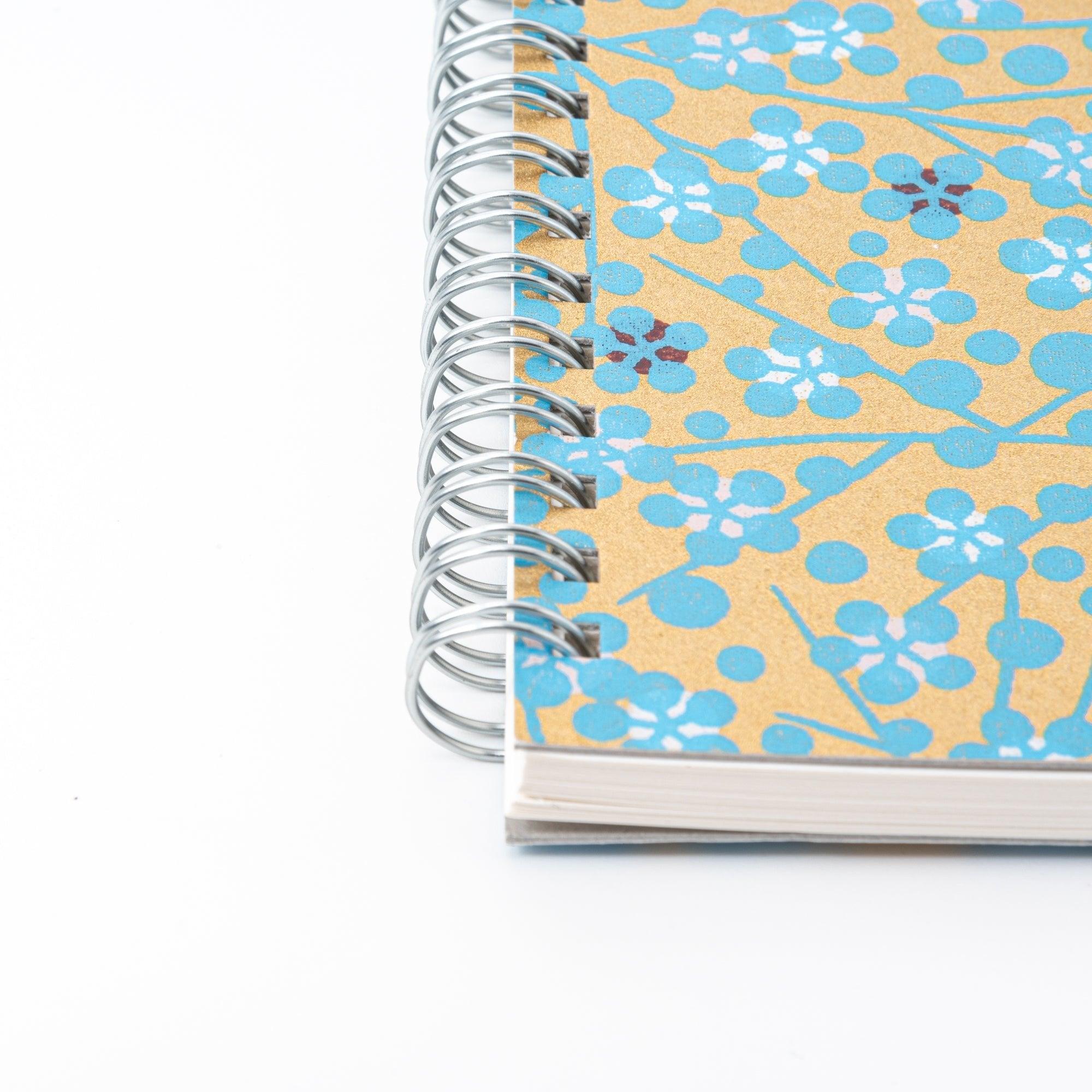 A Japanese notebook with a Yuzen washi cover featuring blue floral patterns on a gold background, displayed on a white desk.