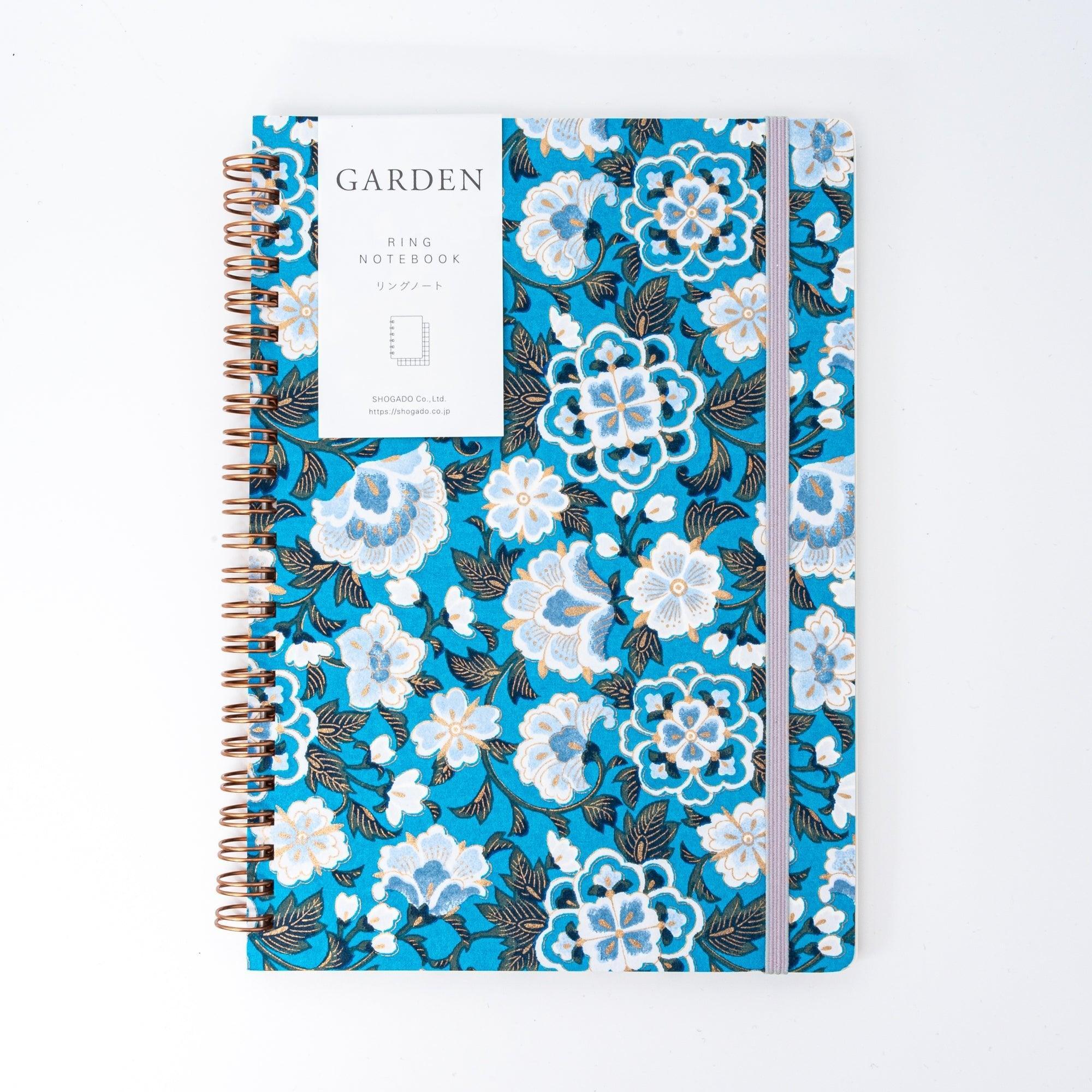 A Japanese notebook with a Yuzen washi cover in vibrant blue, featuring white floral patterns accented by gold, and secured with a light purple elastic band.