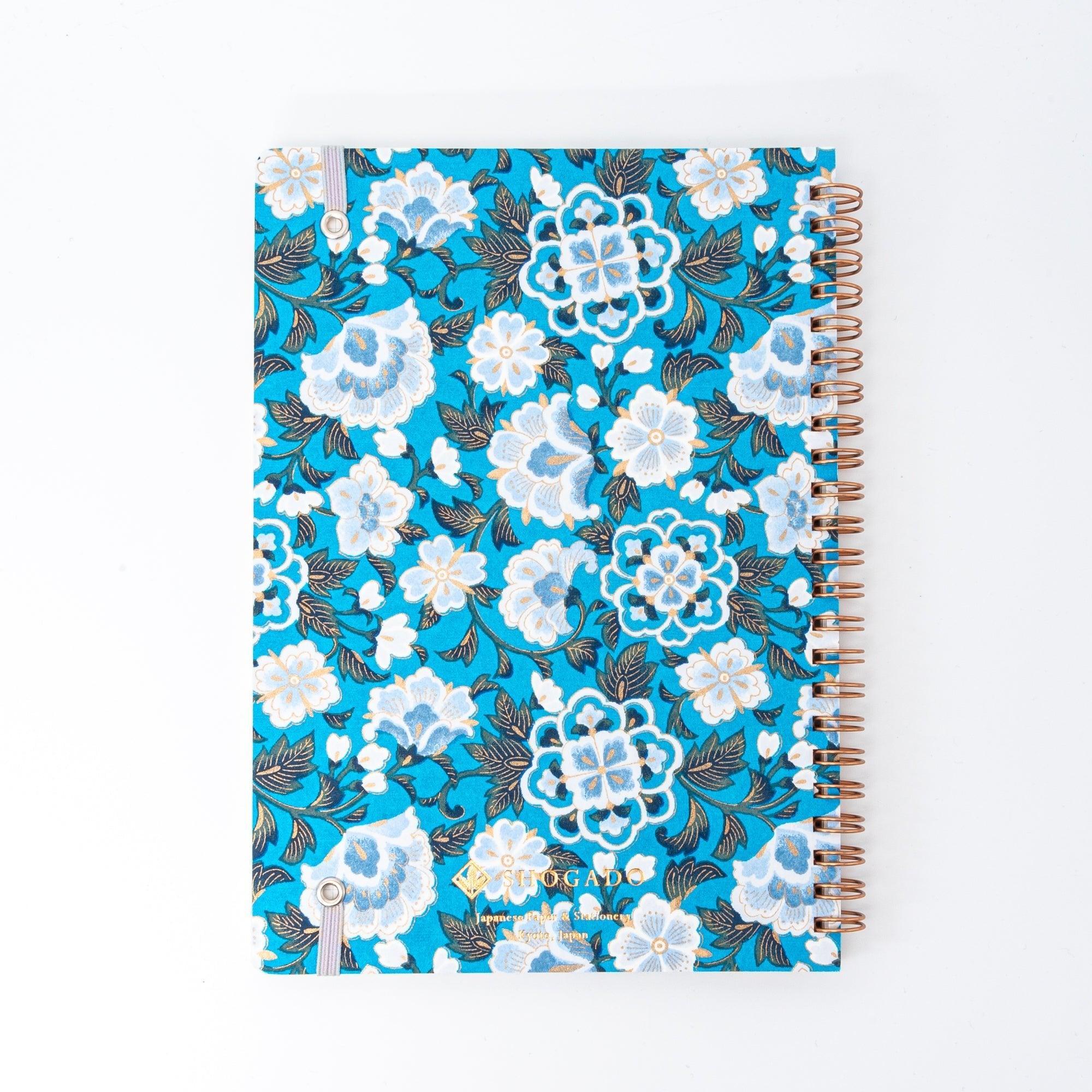 A Japanese notebook with a Yuzen washi cover in vibrant blue, featuring white floral patterns accented by gold, and secured with a light purple elastic band.