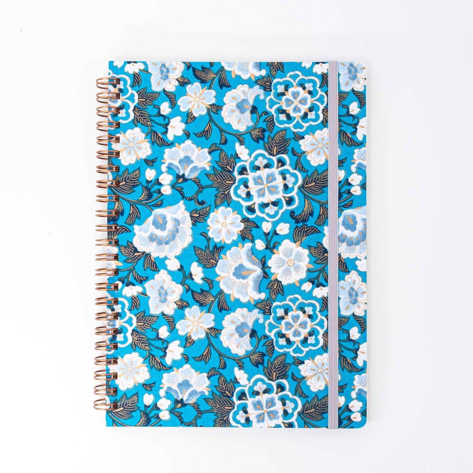A Japanese notebook with a Yuzen washi cover in vibrant blue, featuring white floral patterns accented by gold, and secured with a light purple elastic band.