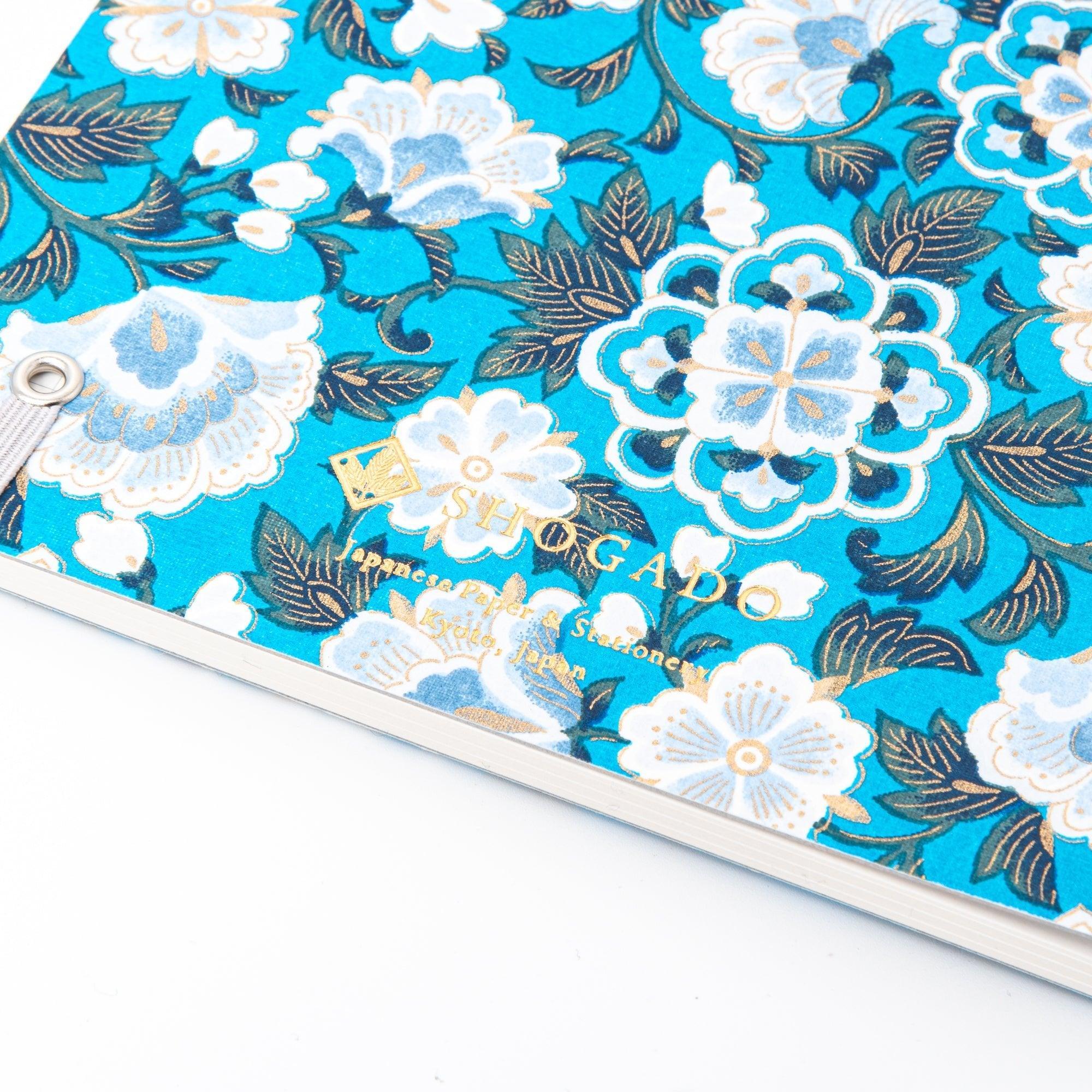 A Japanese notebook with a Yuzen washi cover in vibrant blue, featuring white floral patterns accented by gold, and secured with a light purple elastic band.