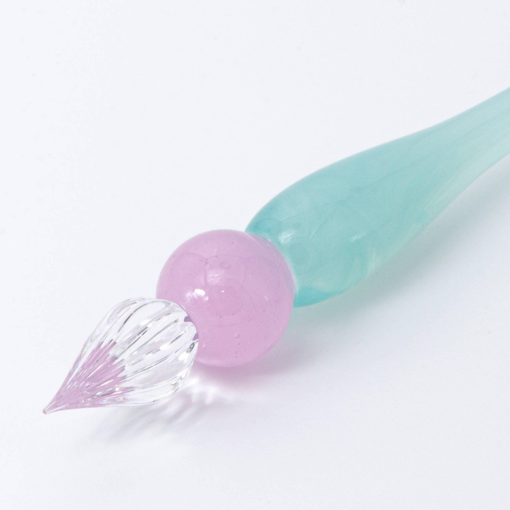 Short glass dip pen with a smooth pastel pink and green gradient body and a clear, spiraled nib, resting on a white surface.