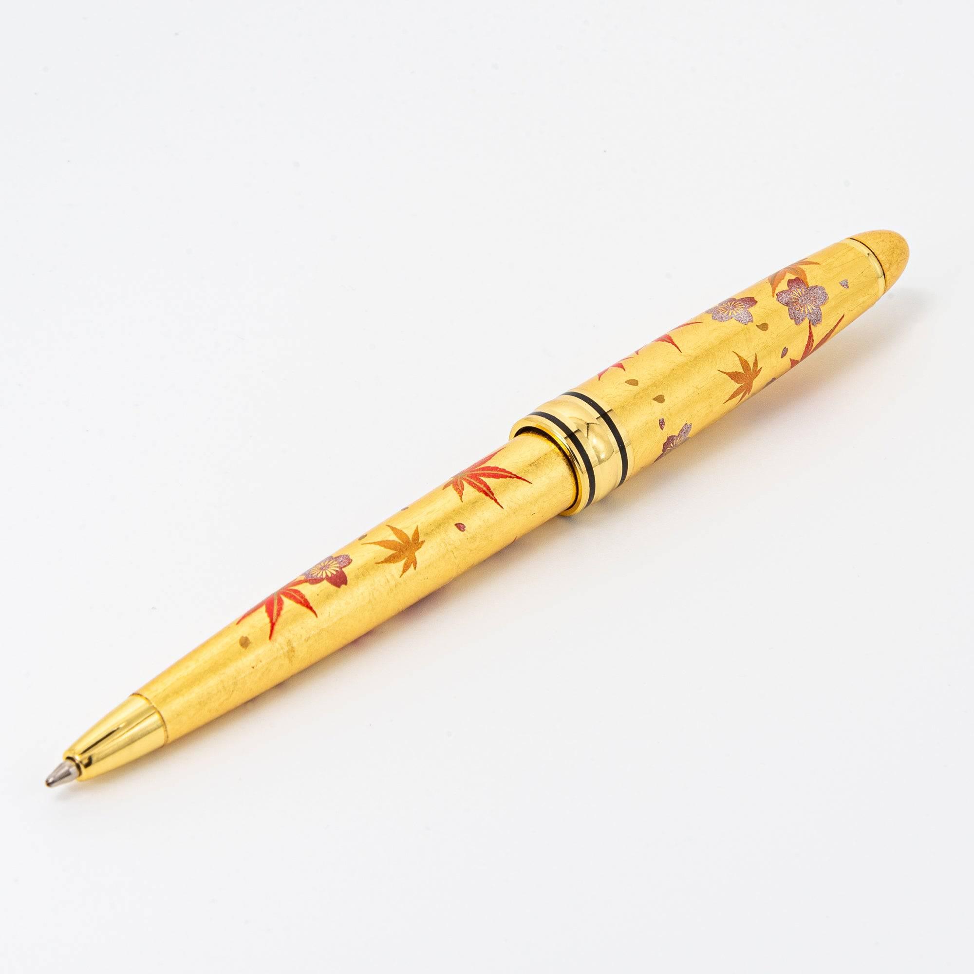A gold ballpoint pen decorated with sakura and momiji leaves, highlighting its lacquered finish and intricate Japanese design.