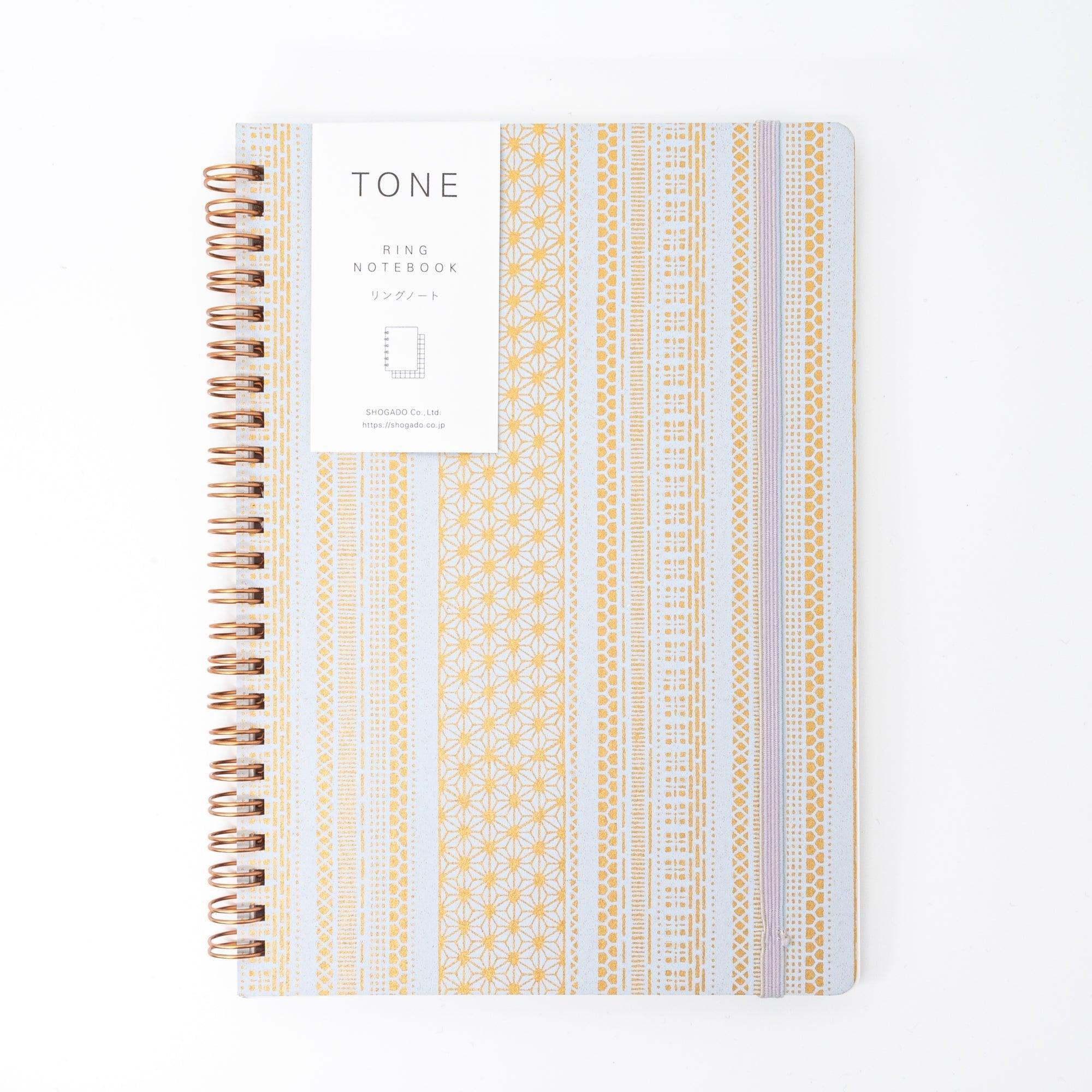 A Japanese notebook with a Yuzen washi cover, featuring yellow and white geometric stripe patterns, rose-gold spiral binding, and an elastic closure band.