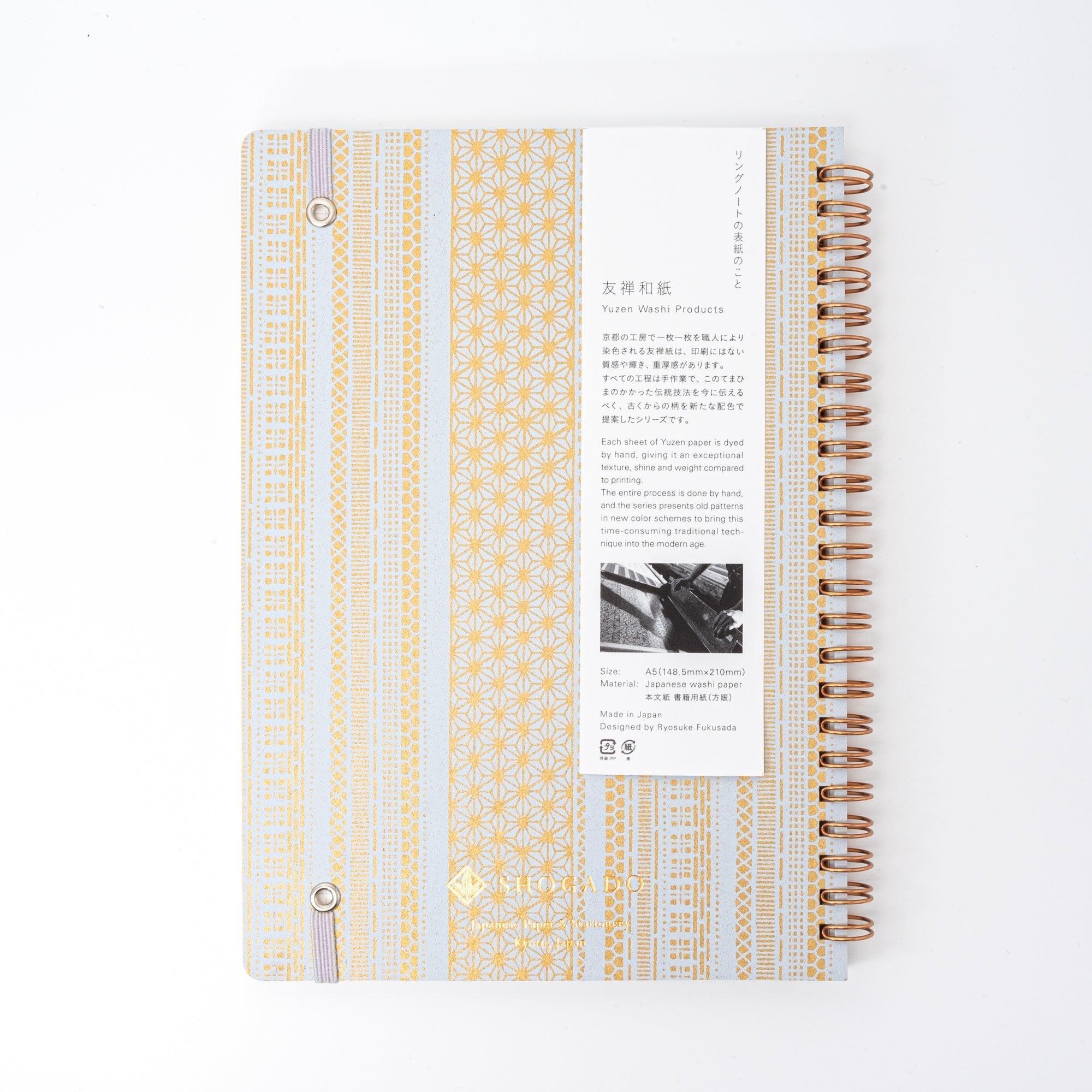 A Japanese notebook with a Yuzen washi cover, featuring yellow and white geometric stripe patterns, rose-gold spiral binding, and an elastic closure band.