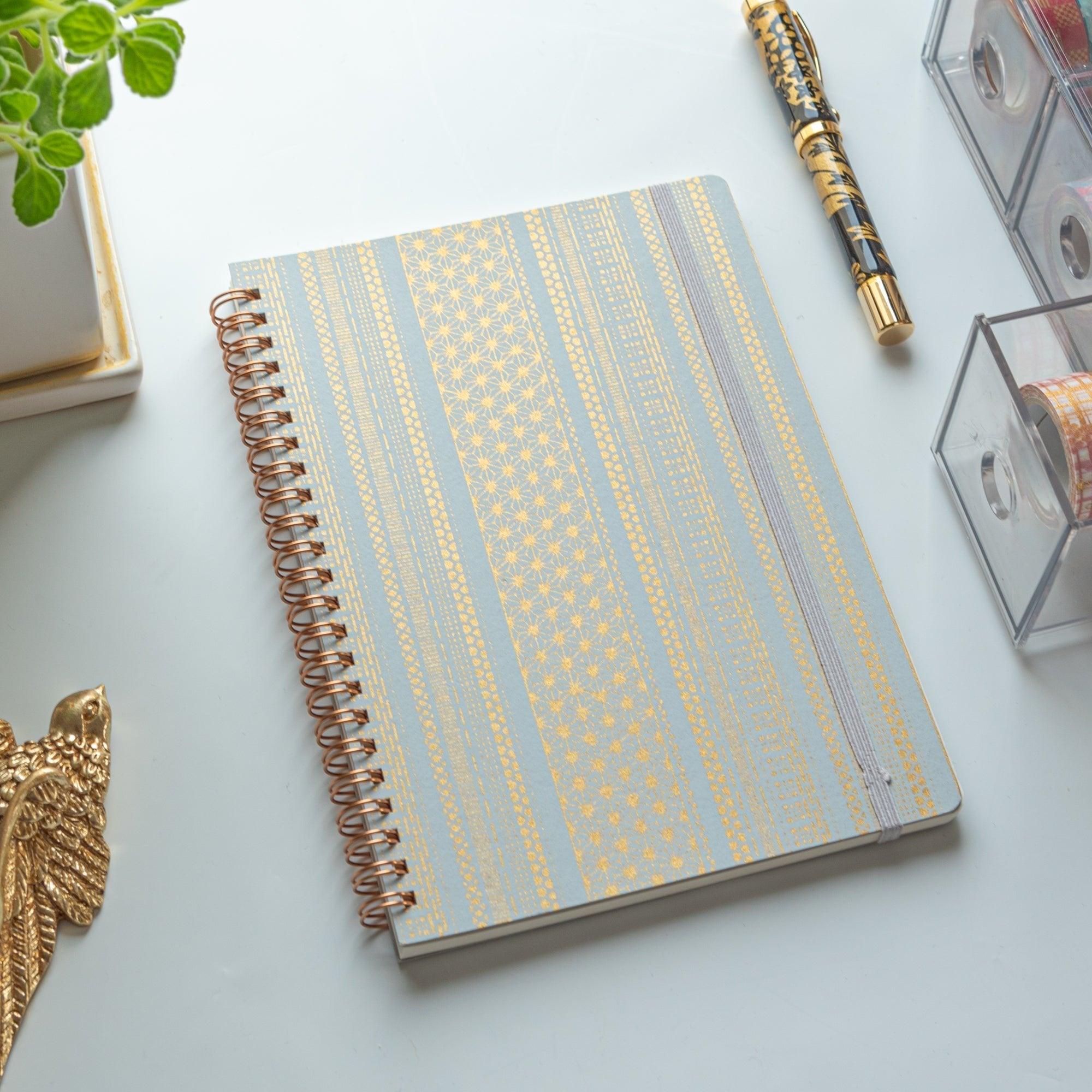 A Japanese notebook with a Yuzen washi cover, featuring yellow and white geometric stripe patterns, rose-gold spiral binding, and an elastic closure band.