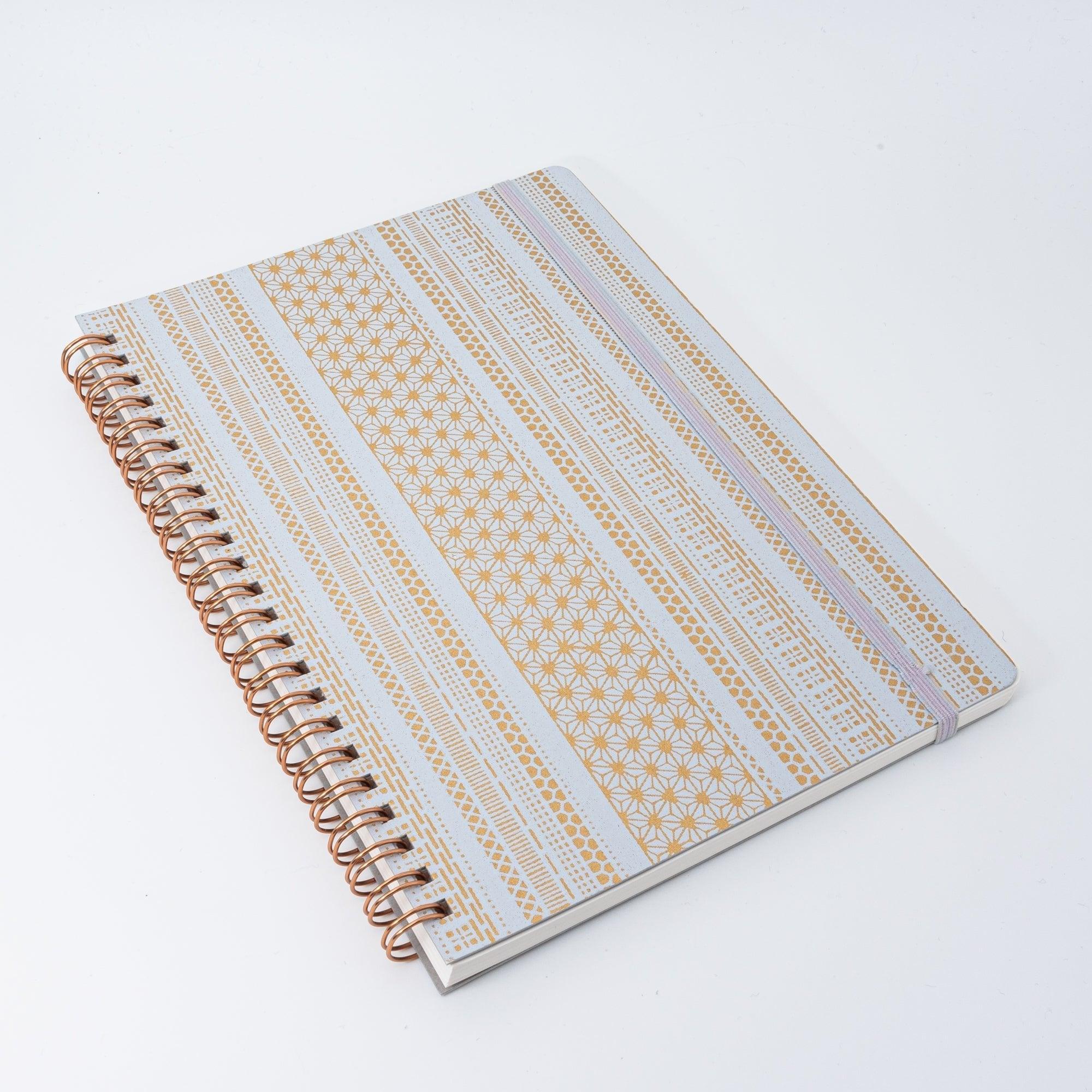 A Japanese notebook with a Yuzen washi cover, featuring yellow and white geometric stripe patterns, rose-gold spiral binding, and an elastic closure band.