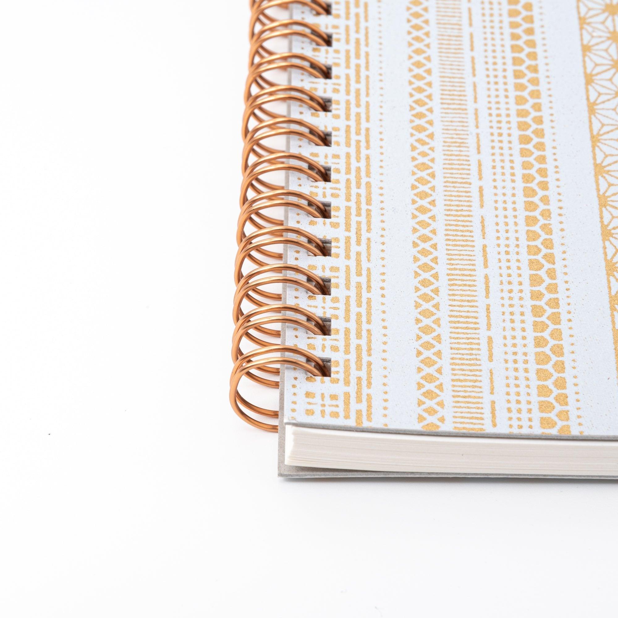 A Japanese notebook with a Yuzen washi cover, featuring yellow and white geometric stripe patterns, rose-gold spiral binding, and an elastic closure band.