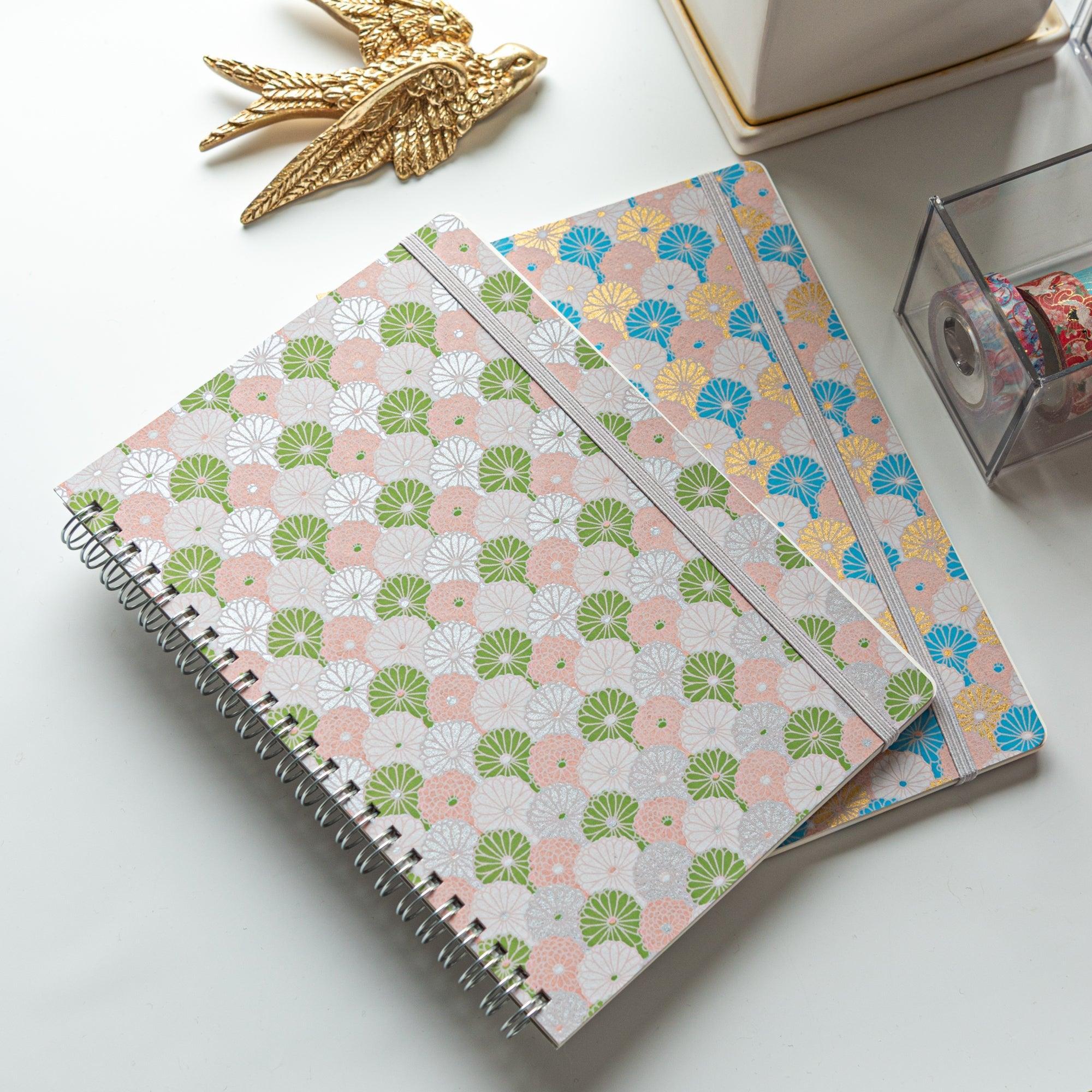 Two Japanese notebooks with Yuzen washi covers featuring pastel fan patterns in green, pink, and blue, placed on a white desk.