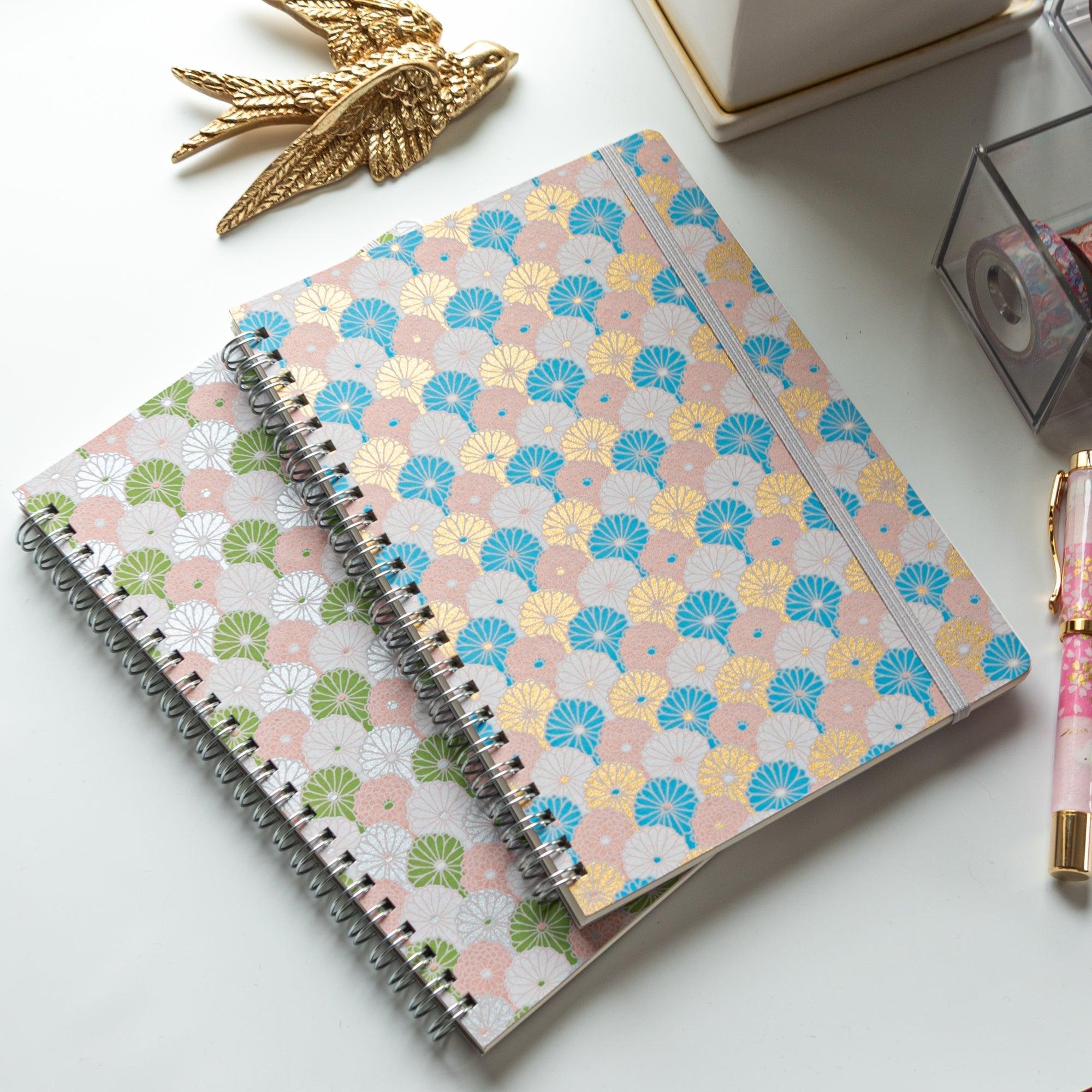 Two Japanese notebooks with Yuzen washi covers featuring pastel fan patterns in green, pink, and blue, placed on a white desk.