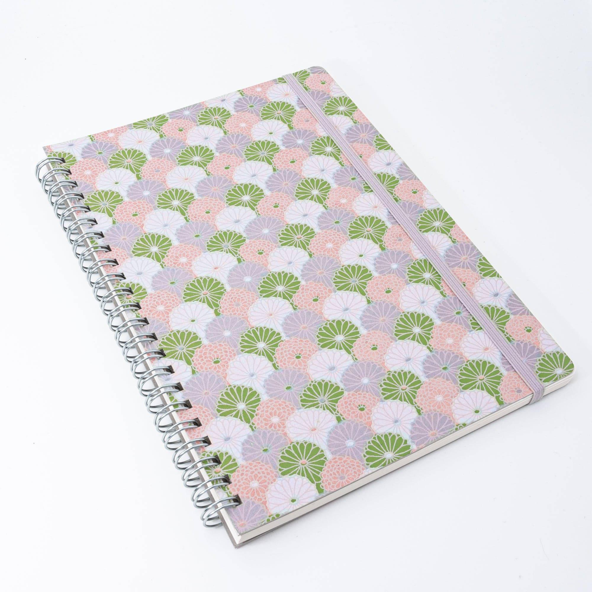 A Japanese notebook with a Yuzen washi cover, showcasing a pastel fan pattern in green, pink, and lilac, with a silver spiral binding and elastic band.