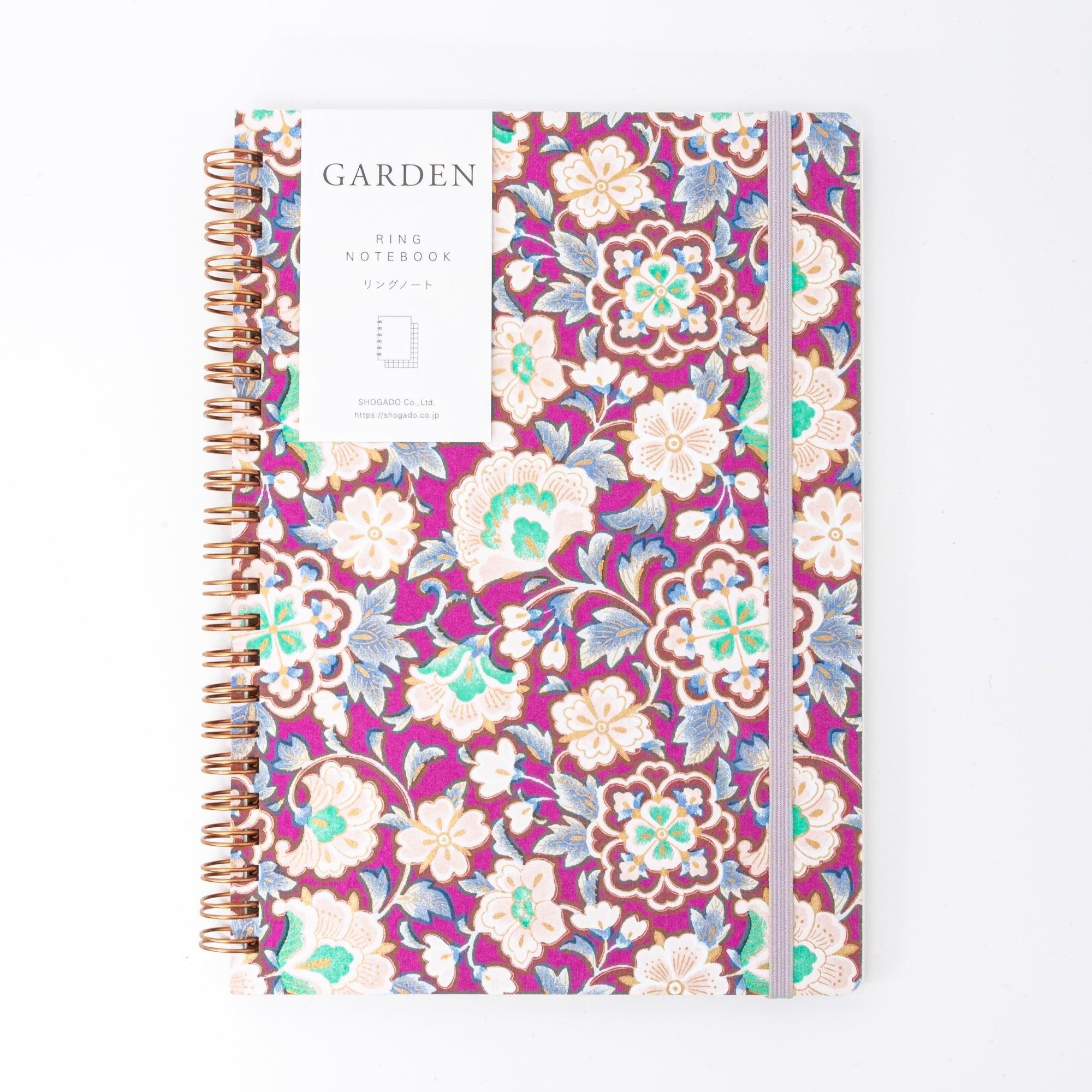 A Japanese notebook with a Yuzen washi cover featuring intricate floral patterns in purple, blue, and green tones, secured with a pink elastic band.
