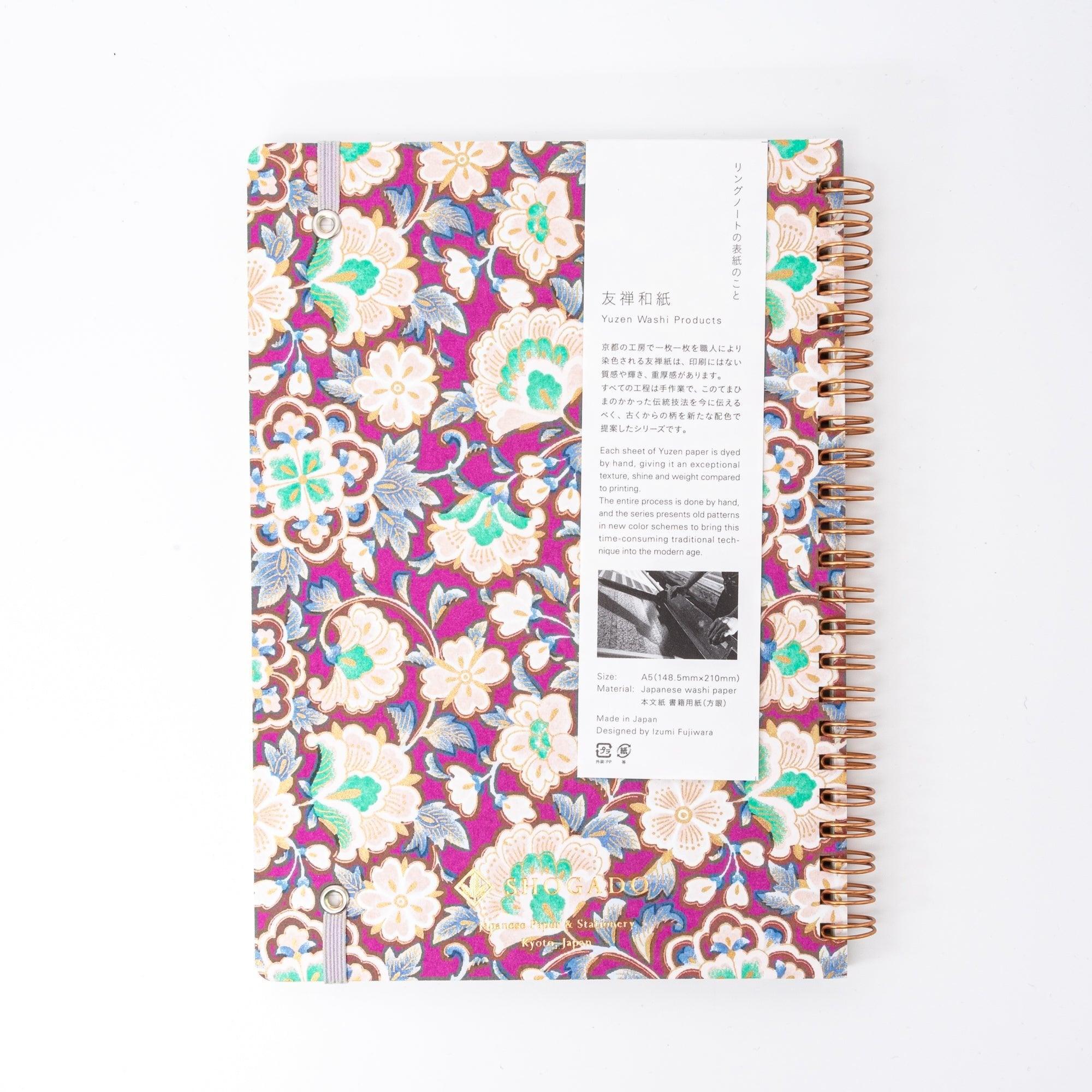 A Japanese notebook with a Yuzen washi cover featuring intricate floral patterns in purple, blue, and green tones, secured with a pink elastic band.