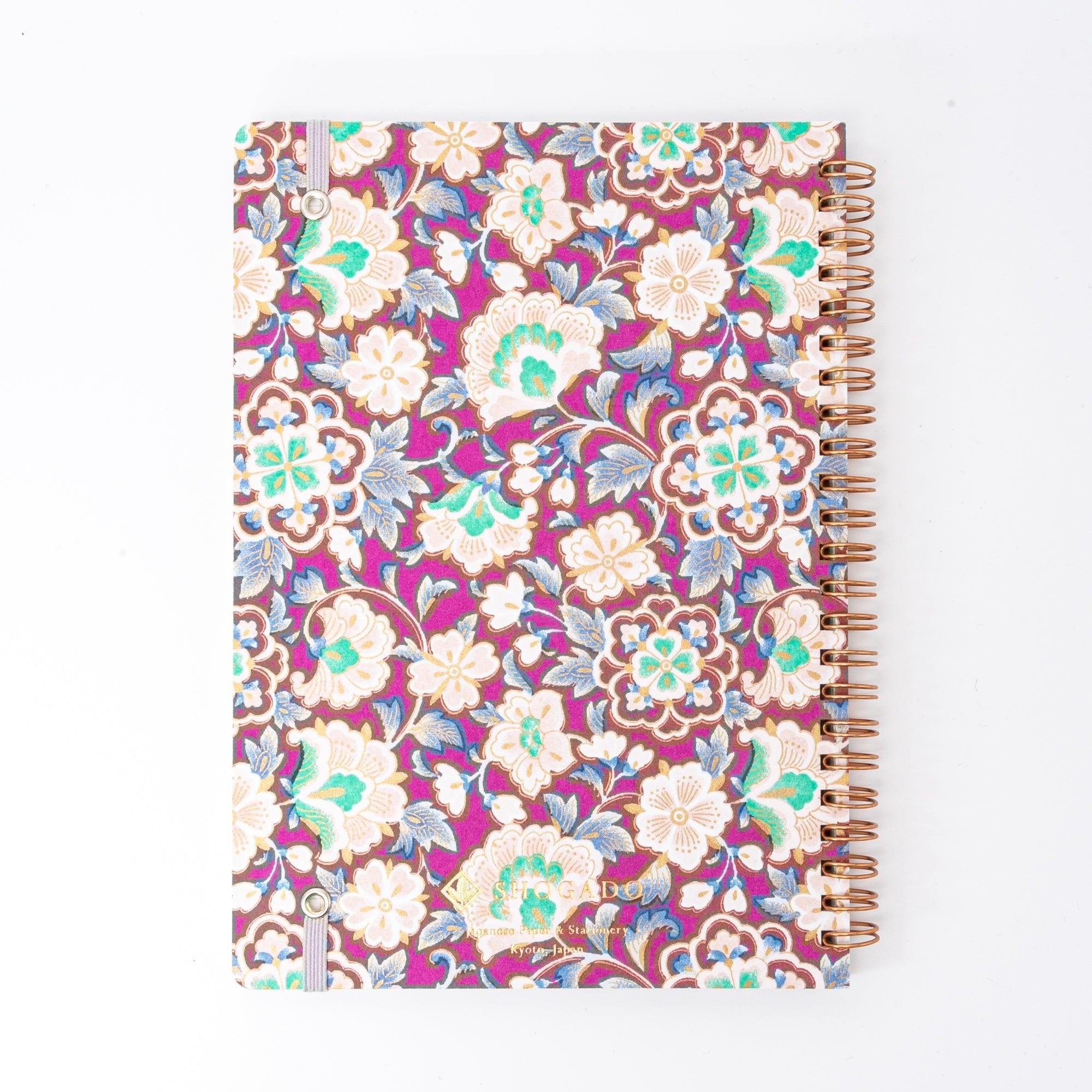 A Japanese notebook with a Yuzen washi cover featuring intricate floral patterns in purple, blue, and green tones, secured with a pink elastic band.