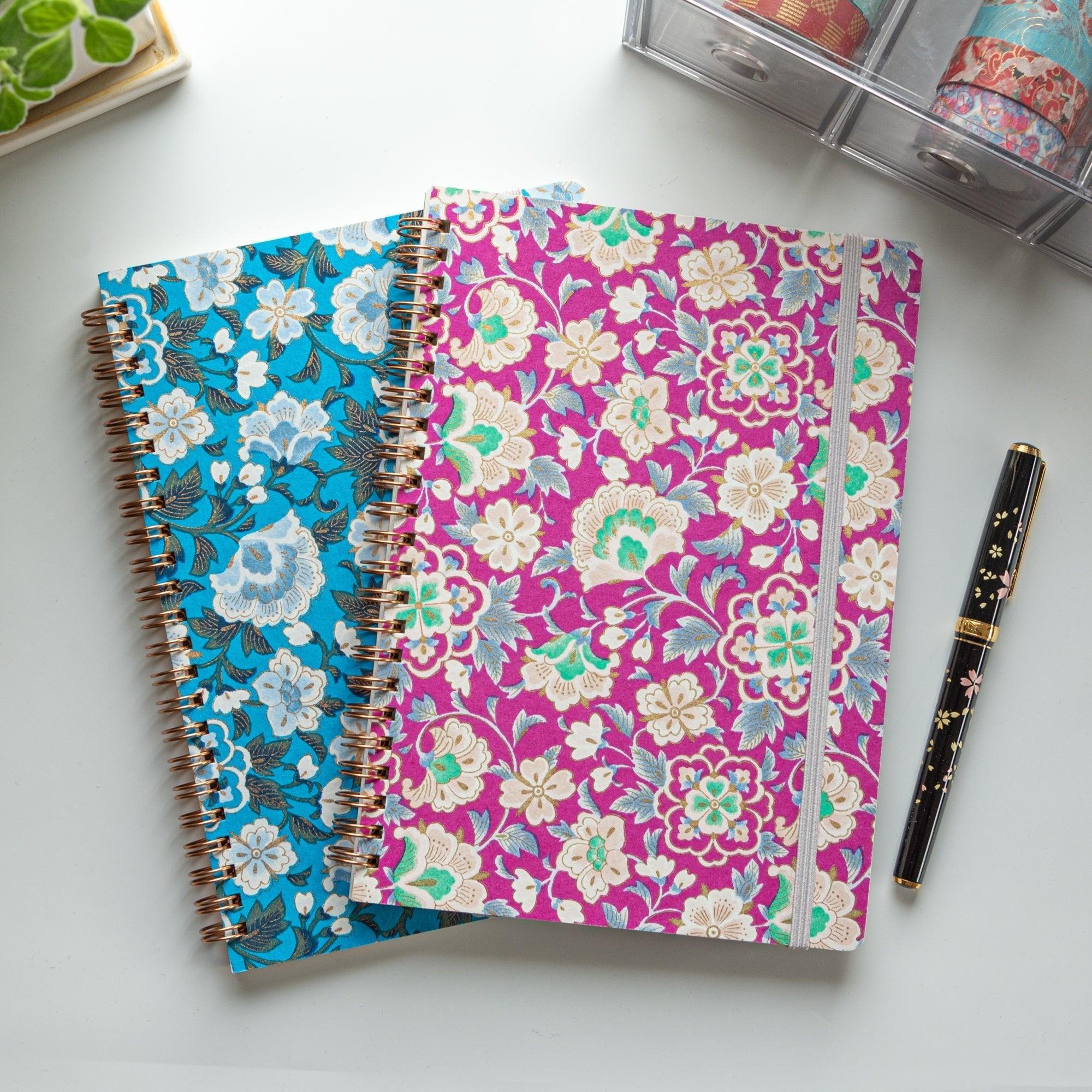 Two Garden Ring Japanese notebooks in magenta and teal, stacked on a desk with a black fountain pen and washi tape organizer