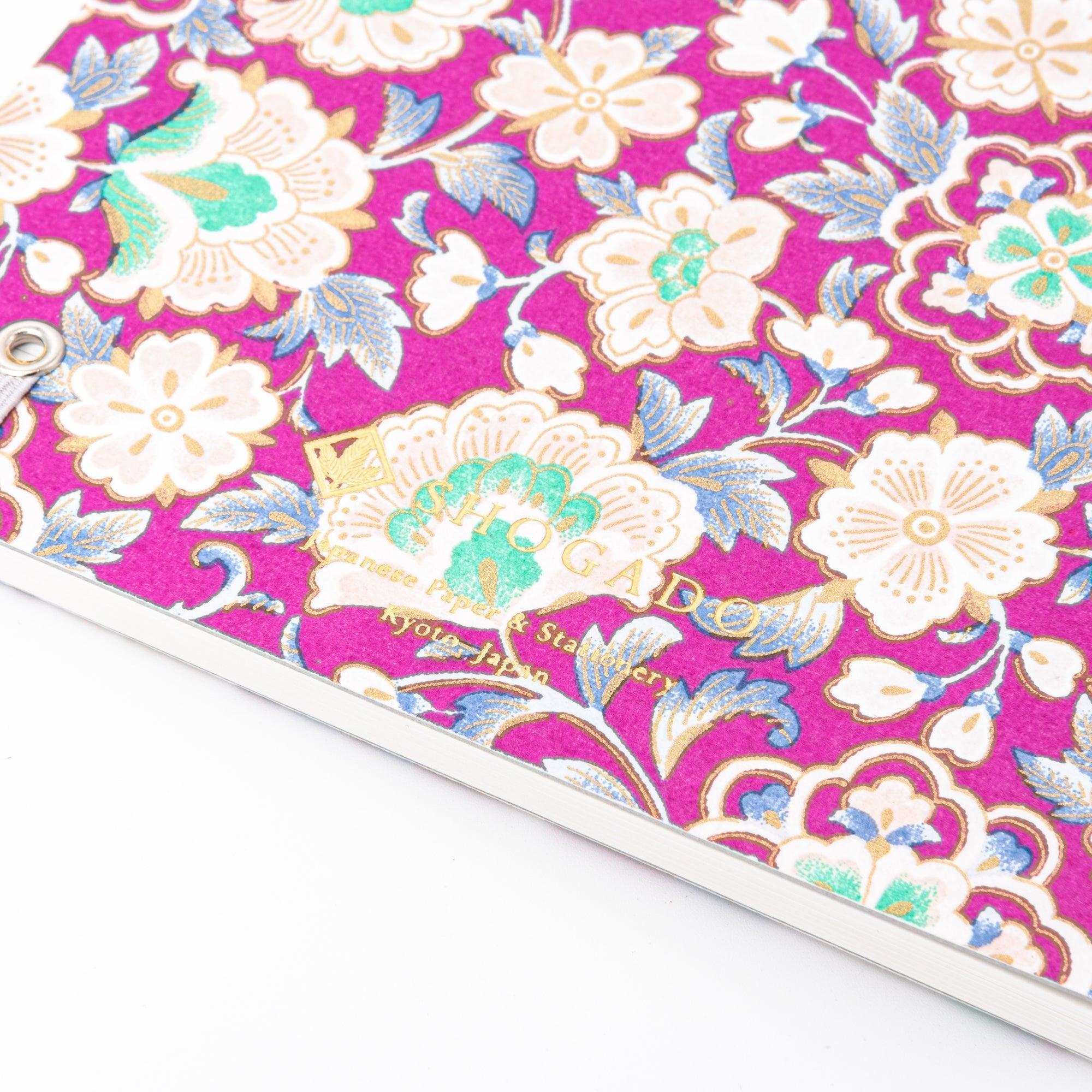 A Japanese notebook with a Yuzen washi cover featuring intricate floral patterns in purple, blue, and green tones, secured with a pink elastic band.