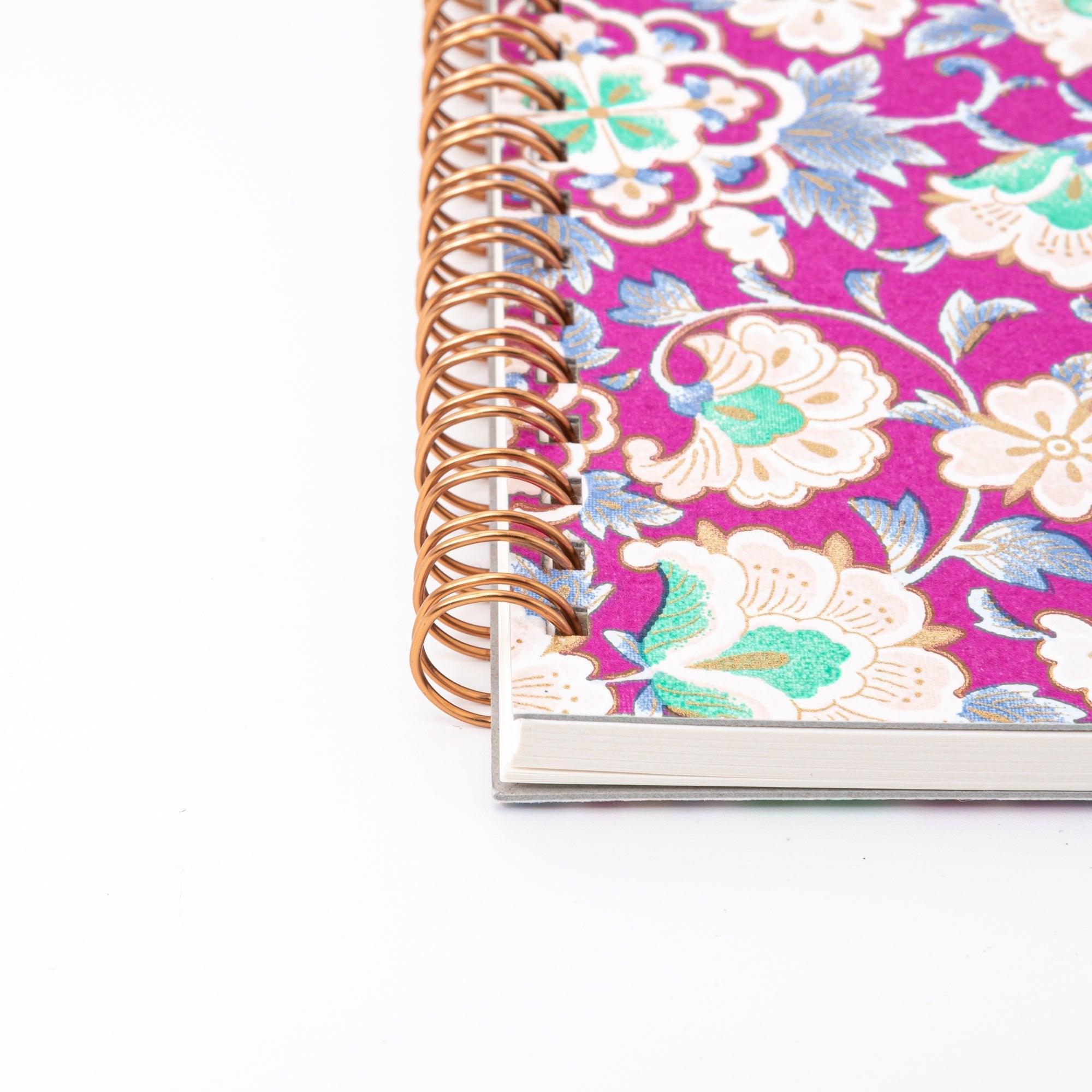 A Japanese notebook with a Yuzen washi cover featuring intricate floral patterns in purple, blue, and green tones, secured with a pink elastic band.