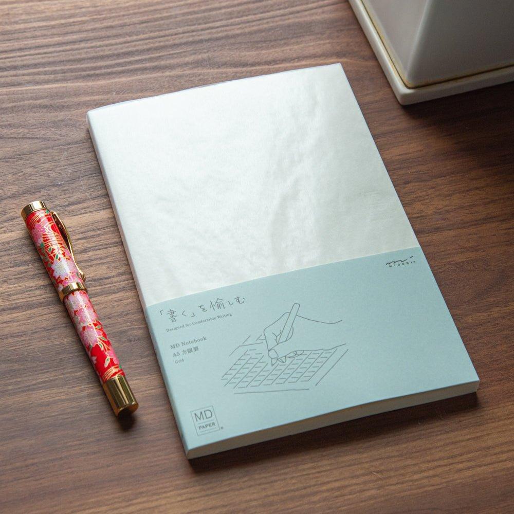 A Japanese stationery MD Notebook with a grid layout is displayed on a wooden desk beside a Japanese fountain pen with a vibrant floral pattern.