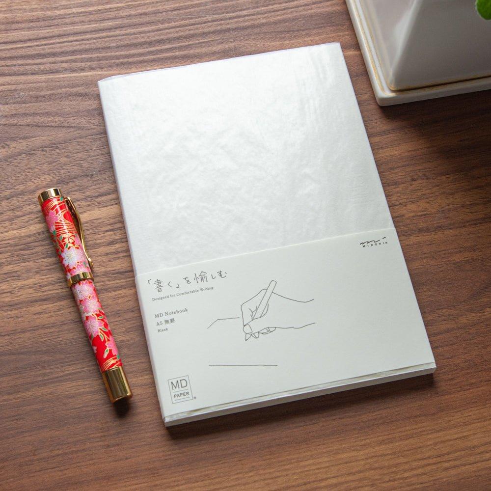 A Japanese stationery MD Notebook with a blank layout is displayed on a wooden desk beside a Japanese fountain pen with floral patterns.