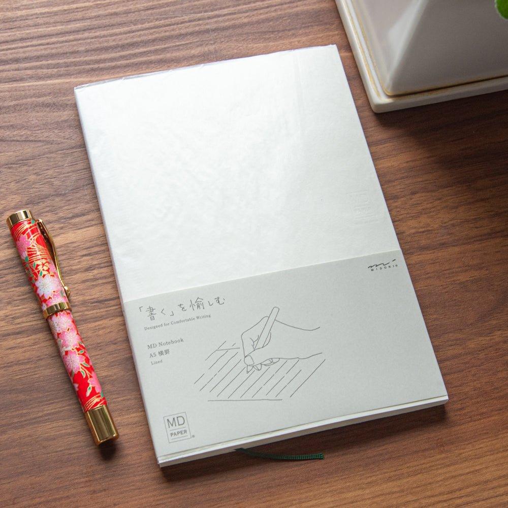 A Japanese stationery MD Notebook placed on a wooden desk beside a colorful Japanese fountain pen. The notebook cover has a simple beige design.