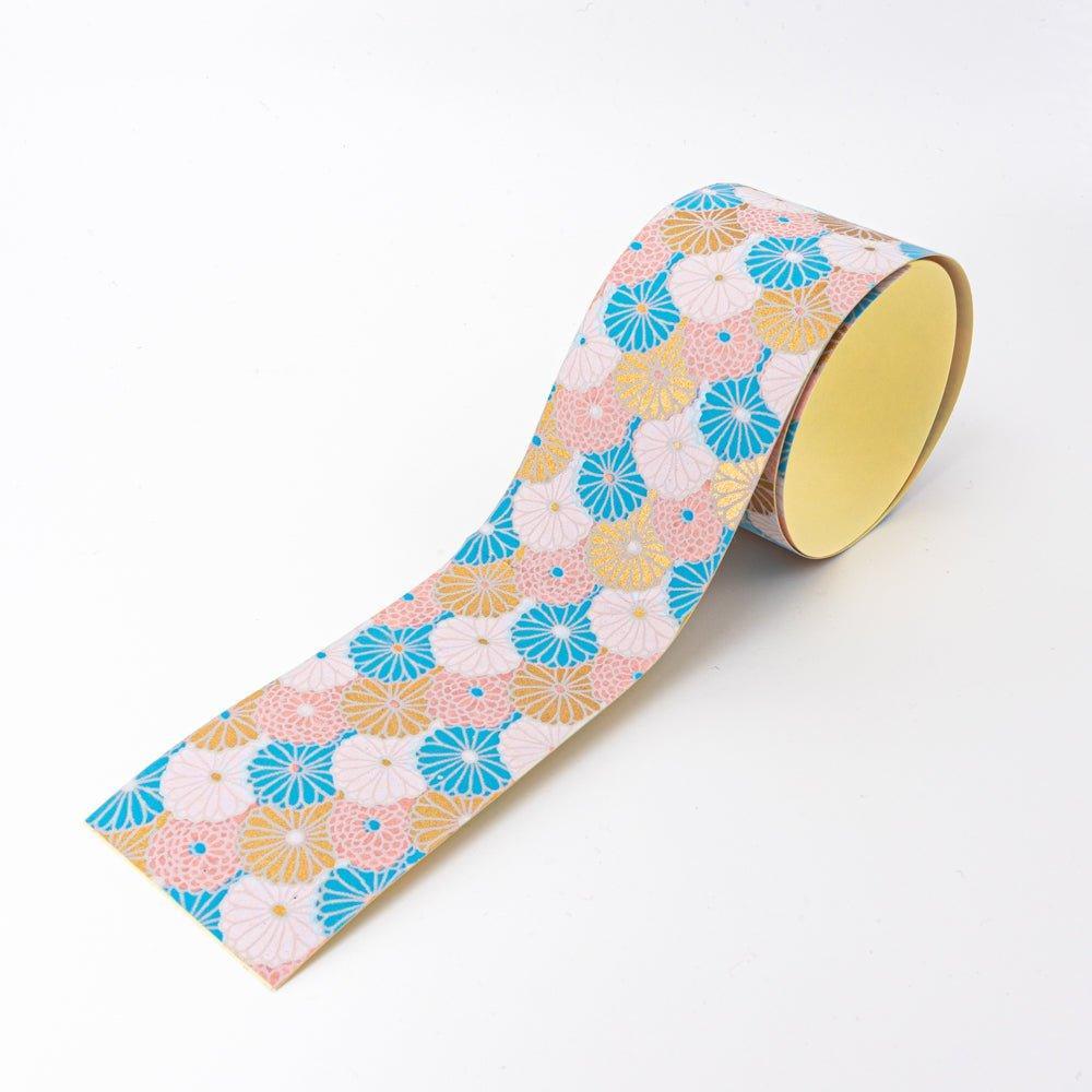 Sky Blossom yuzen washi tape unrolled at angle showing chrysanthemum in blue, pink, and gold on white, yellow backing visible
