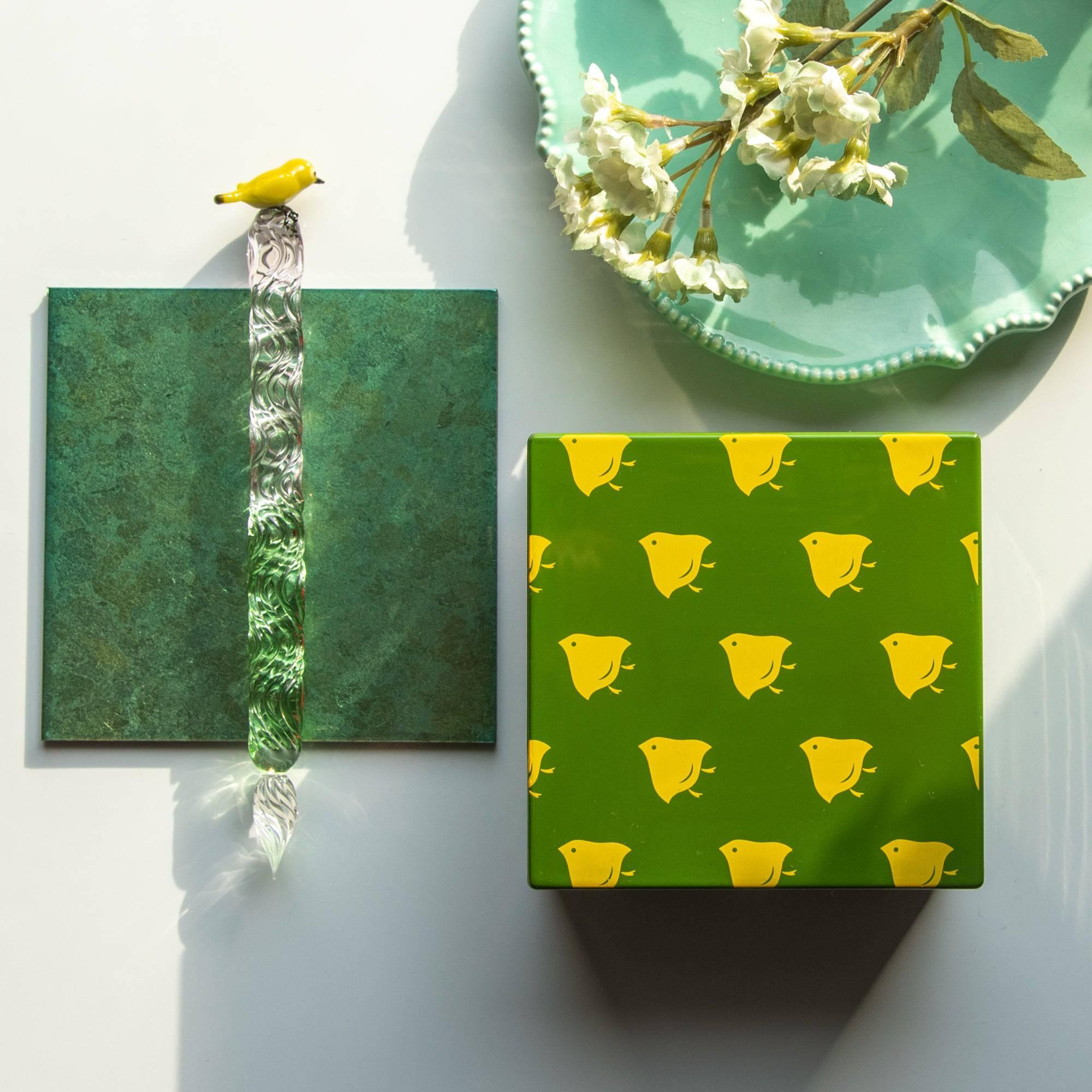 Green Japanese lacquer stationery box with yellow bird patterns, accompanied by a green glass pen, floral plate, and greenery for an artistic desk arrangement.