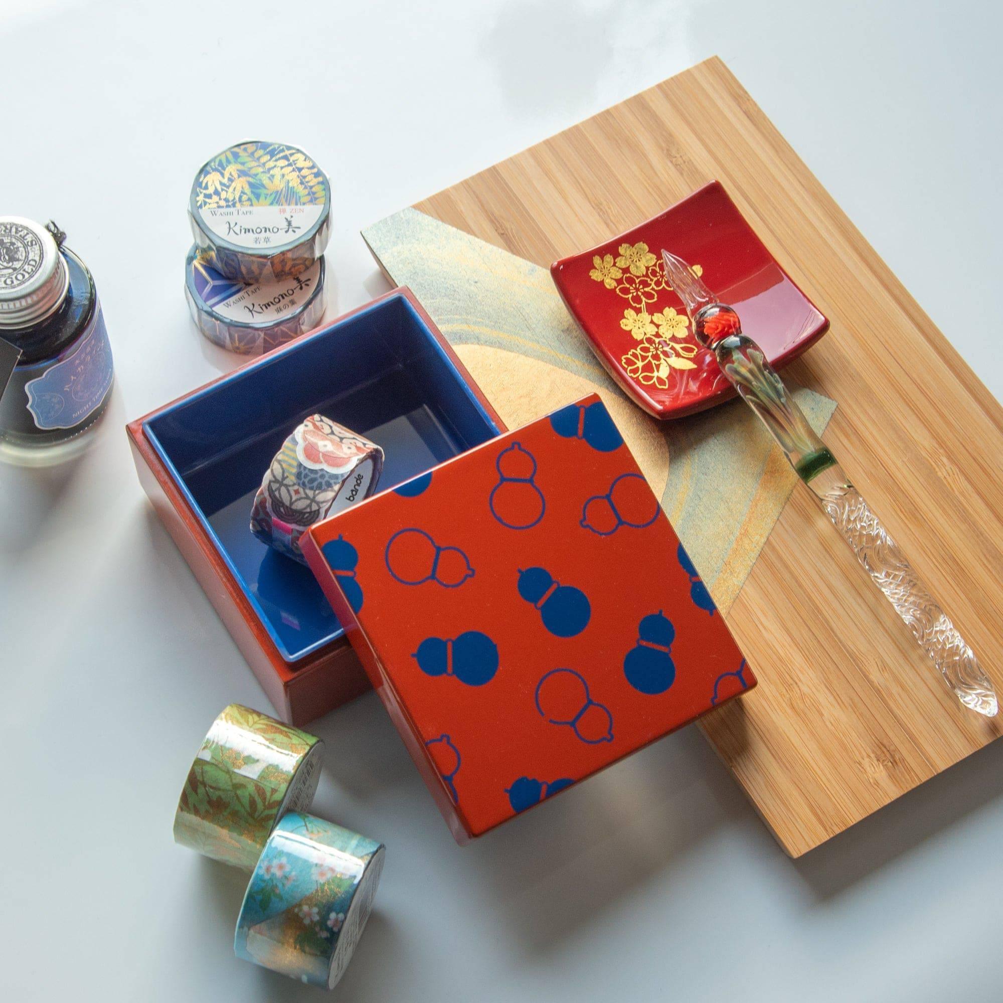 Open red Japanese lacquer stationery box with blue gourd patterns, containing washi tape rolls, alongside a decorative tray and glass pen on a desk.