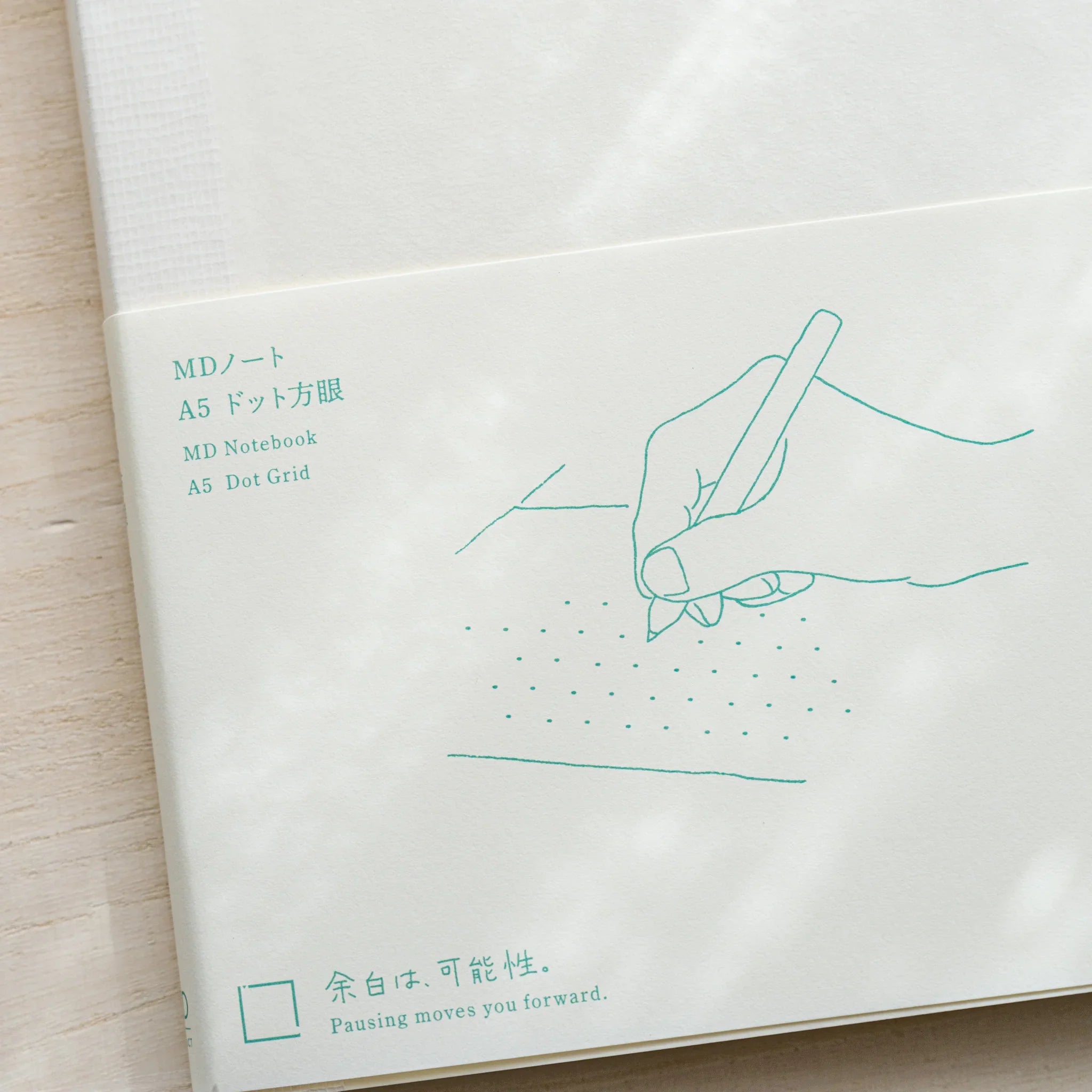Midori MD Notebook A5 Dot Grid belly band with teal line illustration of hand holding pen, text MD Notebook A5 Dot Grid
