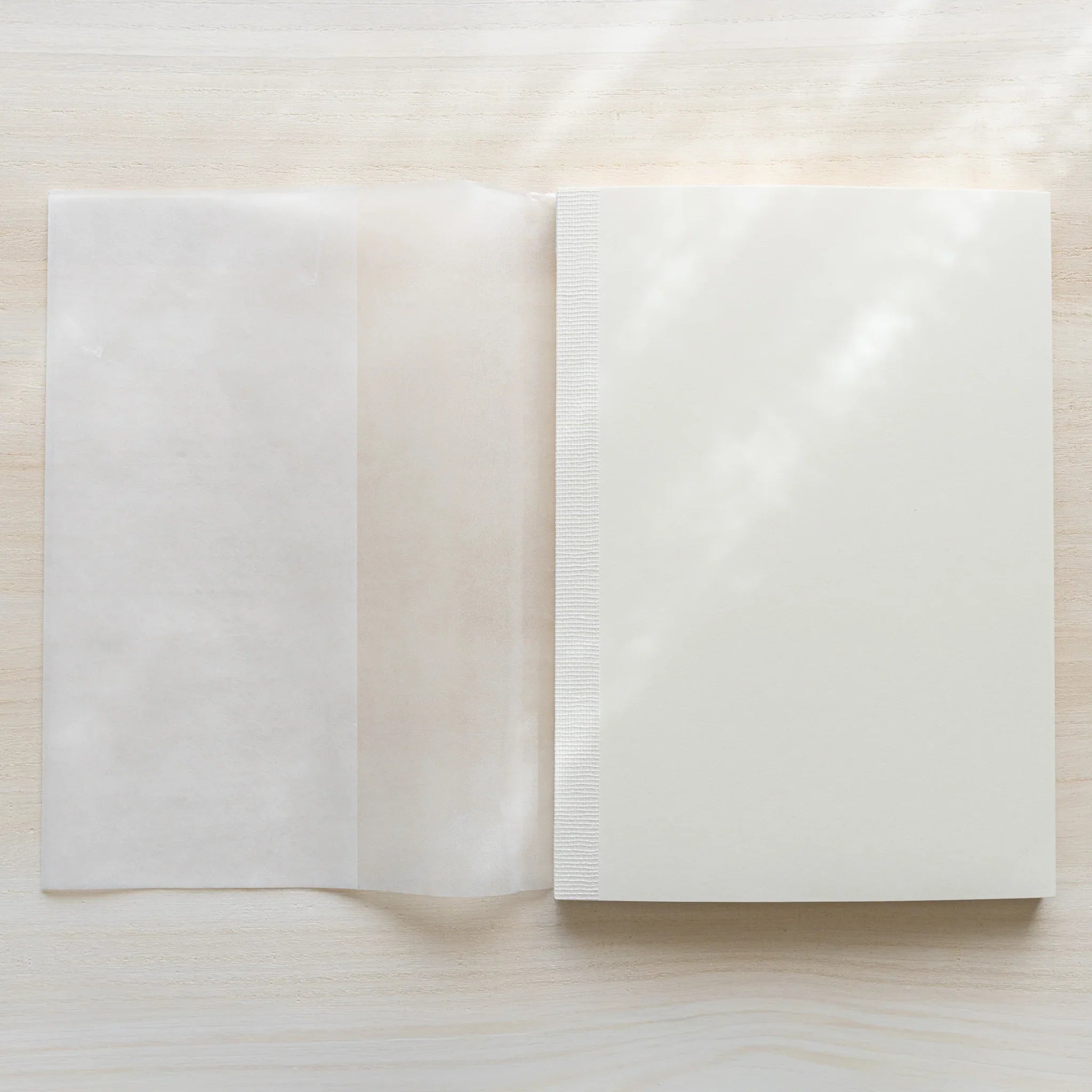 Midori MD Notebook A5 next to unfolded glassine wrapping on light wood, showing white linen cover and thread-stitched spine
