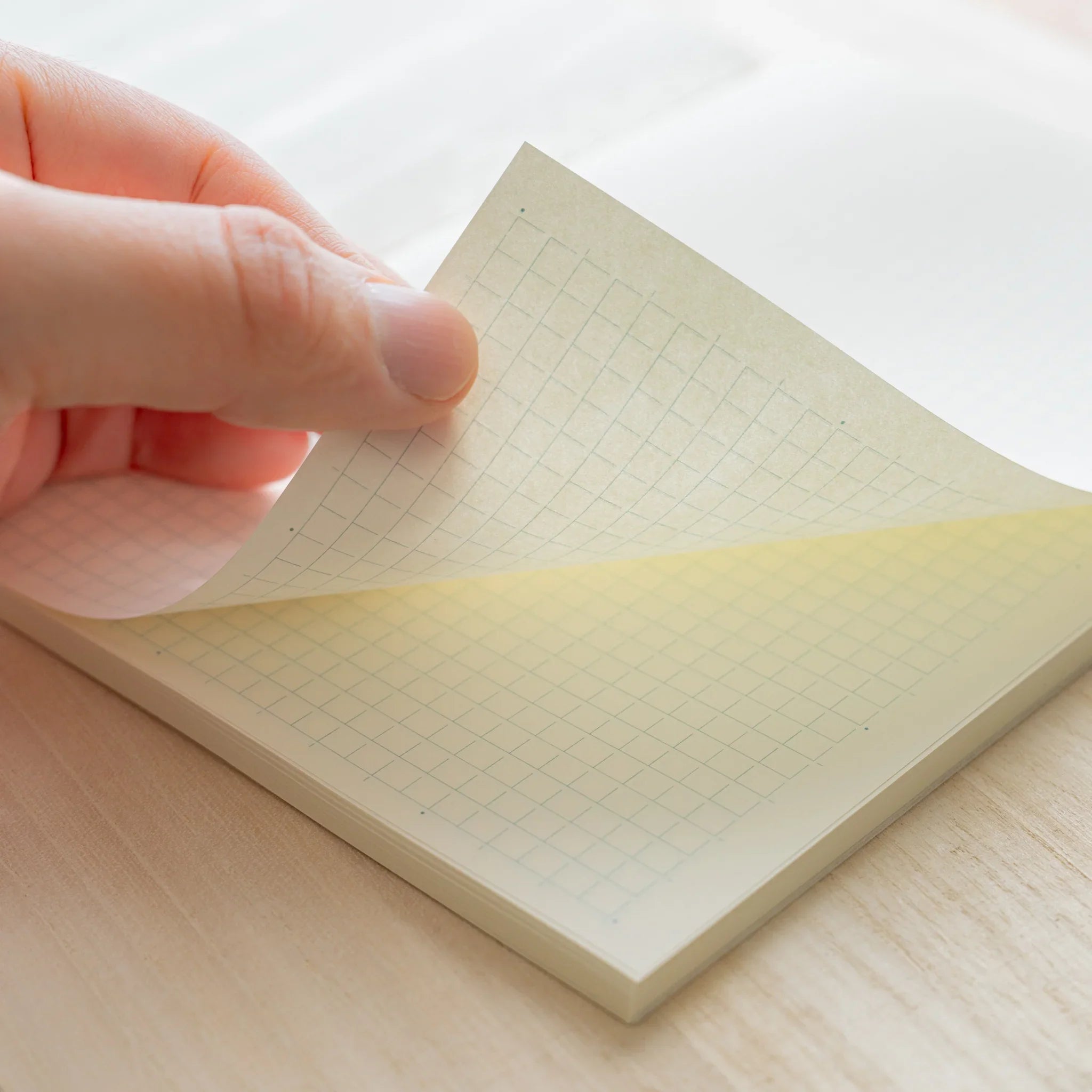 Hand turning ivory grid page of Midori MD Notebook A5, showing warm ivory paper tone, grid lines and page thickness on wood