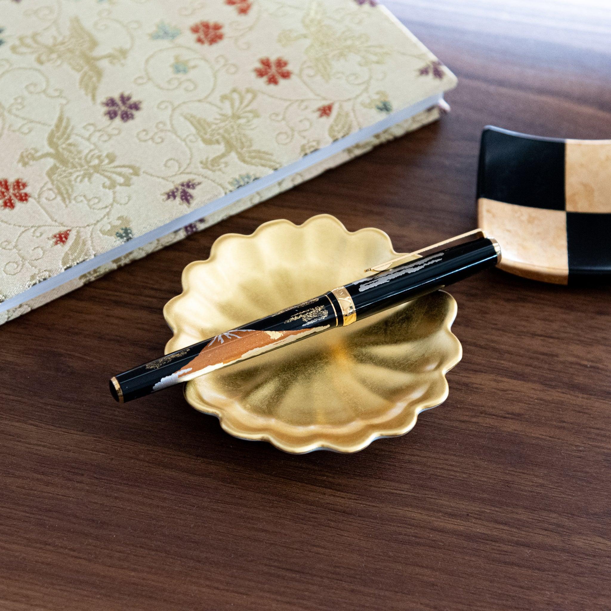 A Japanese stationery fountain pen on a gold scalloped dish, alongside a black and gold checkered square tray and floral brocade notebook.  