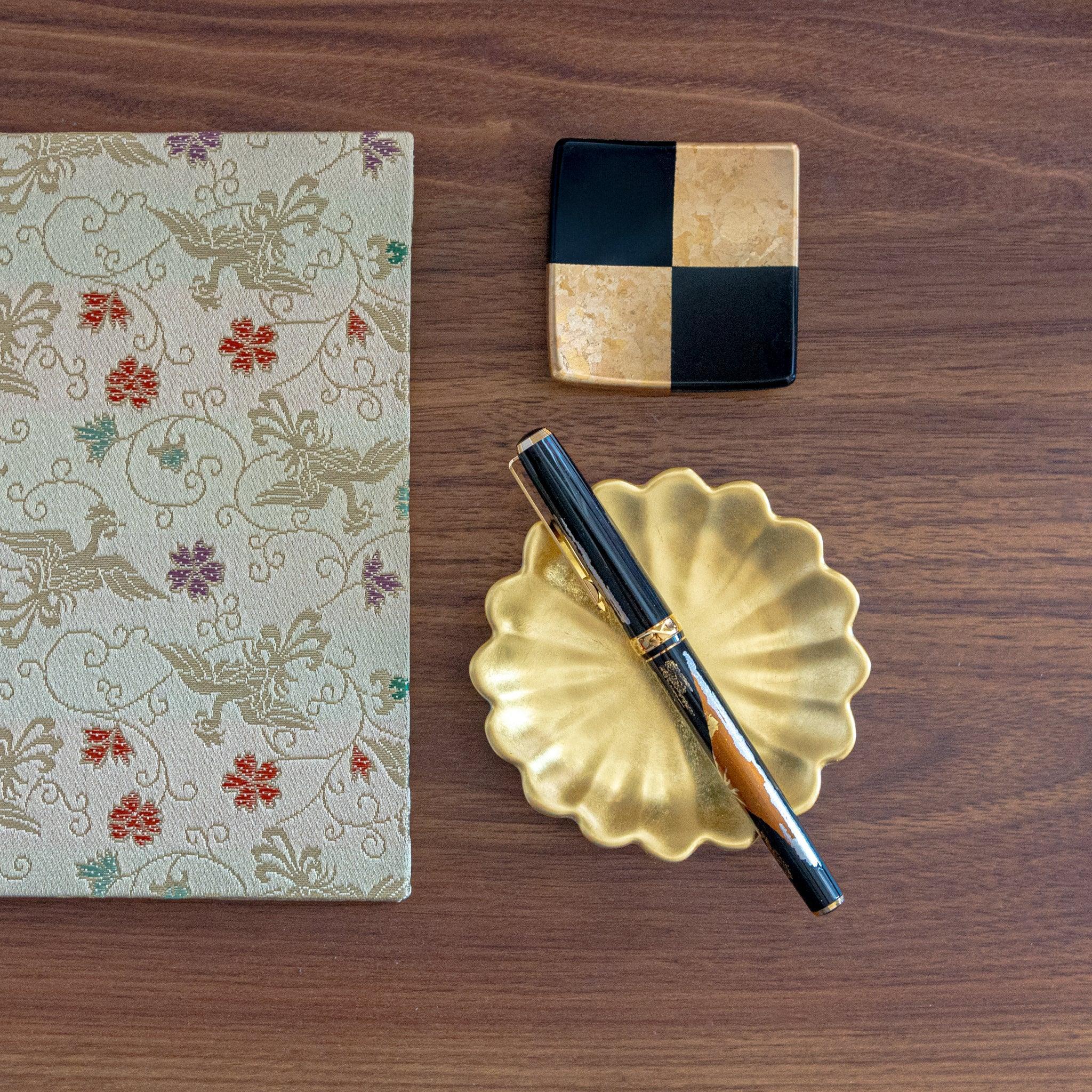 A Japanese stationery fountain pen on a gold scalloped dish, alongside a black and gold checkered square tray and floral brocade notebook.  