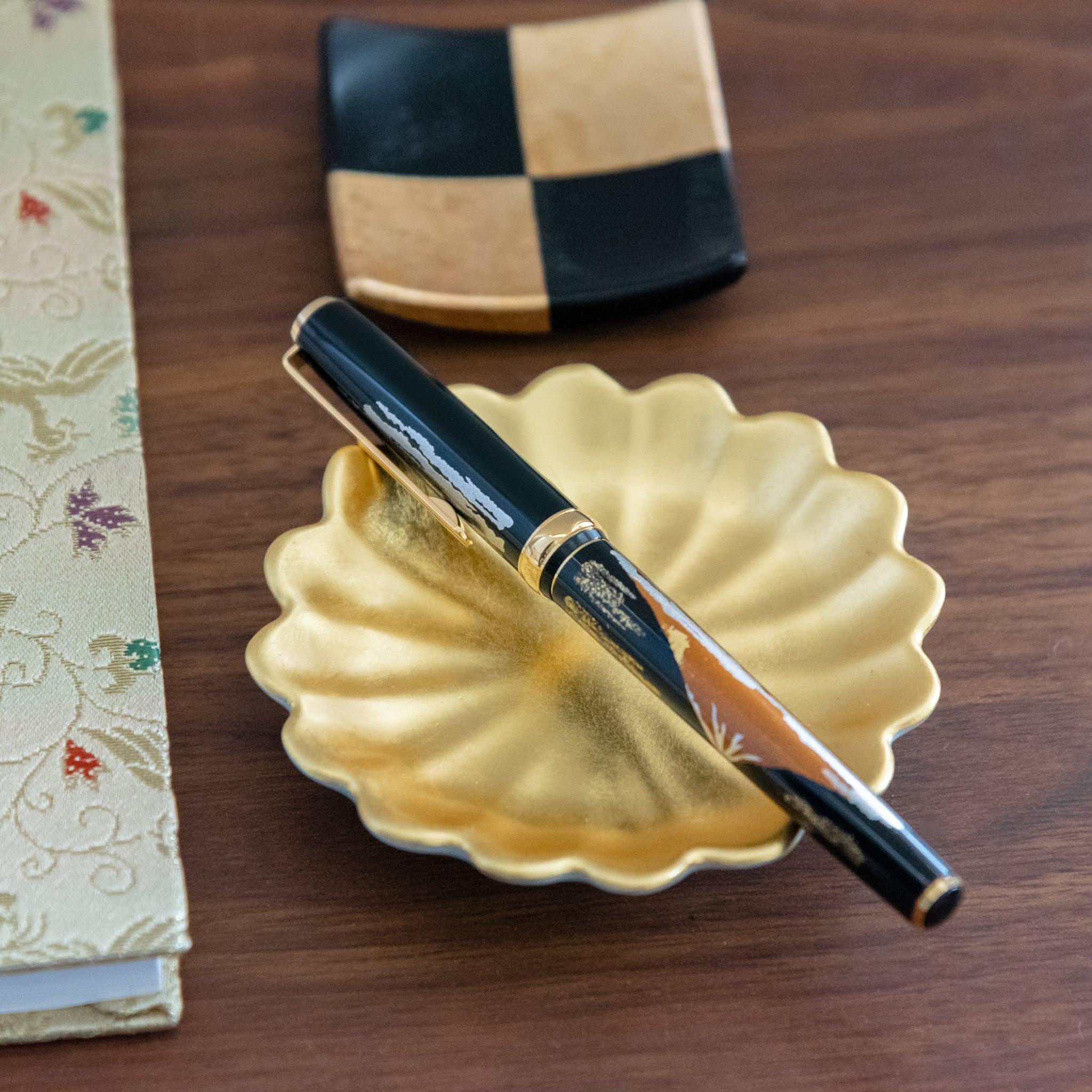 A Japanese stationery fountain pen with a Mt. Fuji design, resting on a gold dish, paired with a checkered black and gold tray and brocade notebook.  