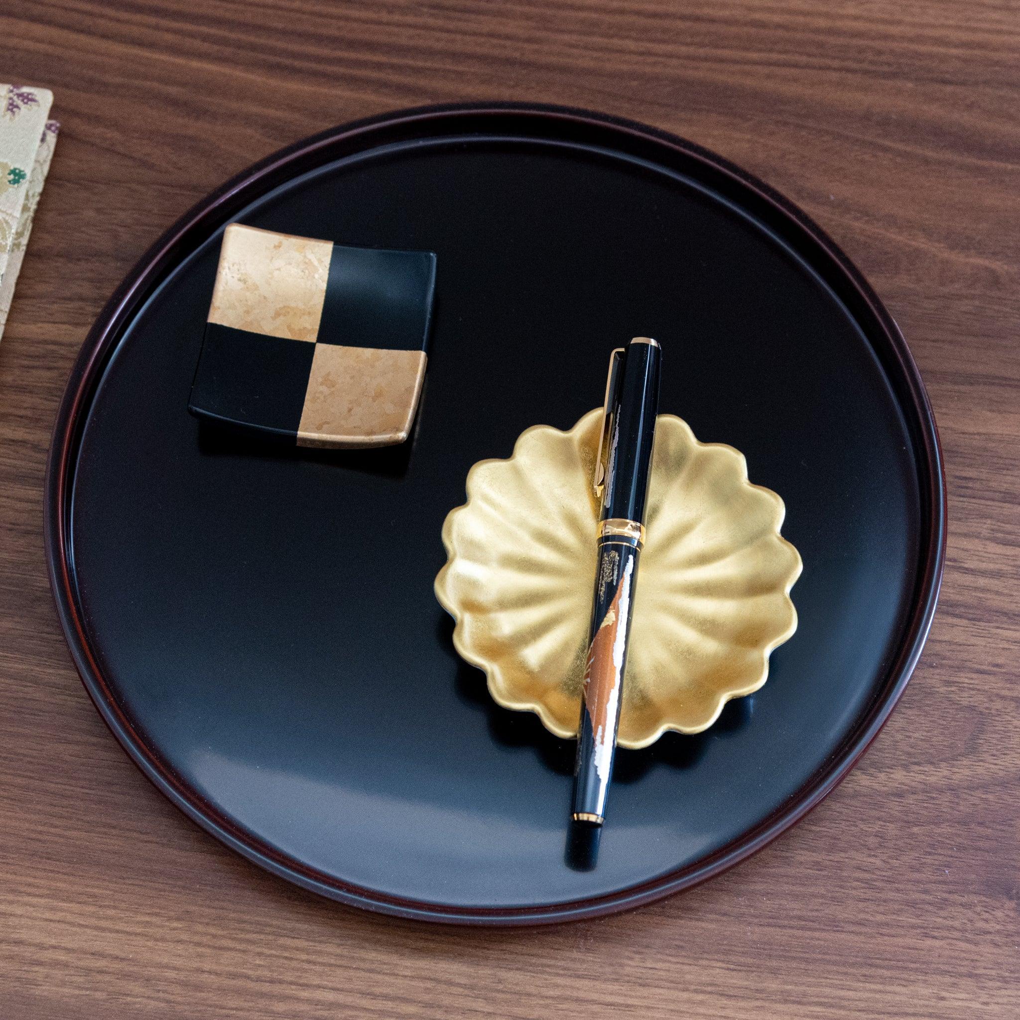 A Mt. Fuji-themed Japanese stationery fountain pen displayed on a gold dish, placed on a black lacquer tray with a checkered accent piece.  