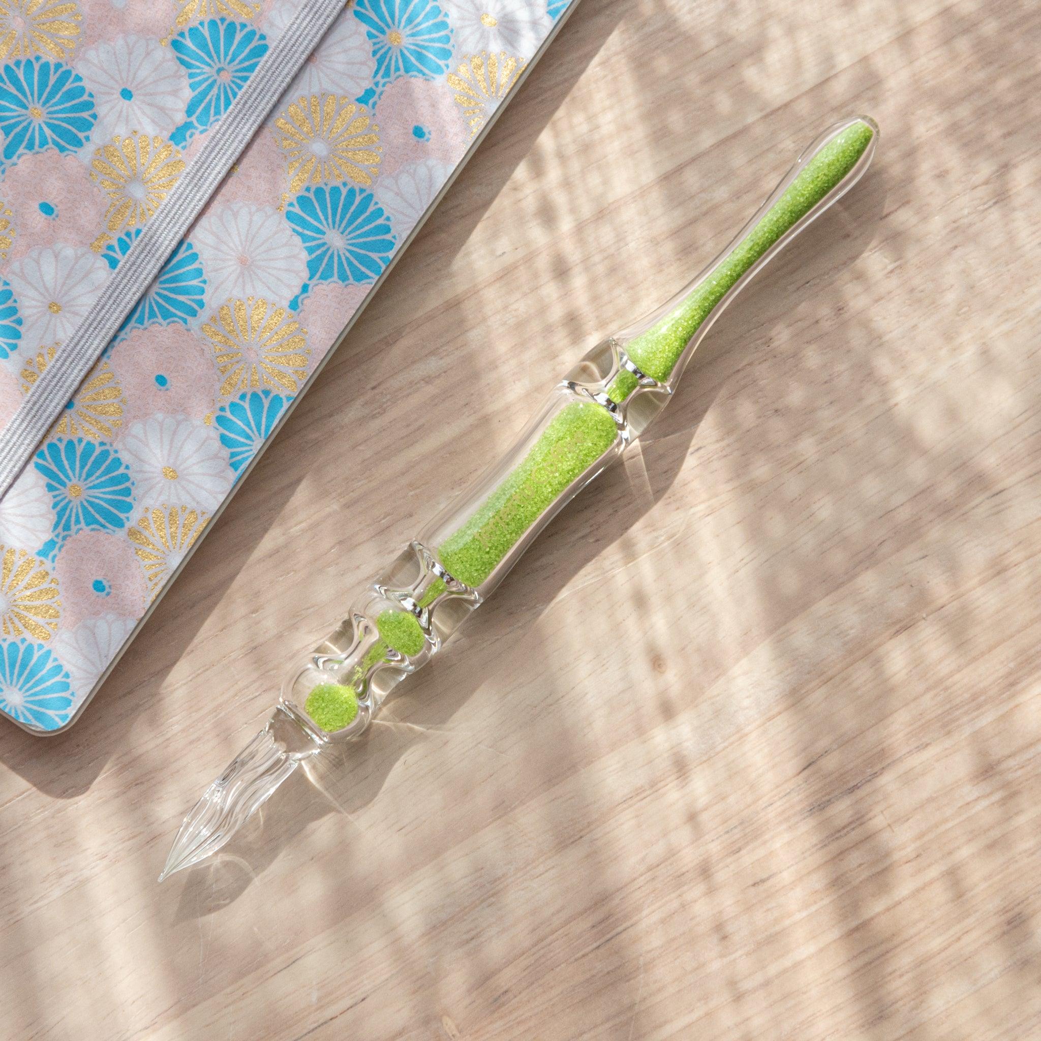 A glass dip pen with green sand-like particles inside its shaft rests on a wooden surface next to a pastel floral-patterned notebook. The pen's transparent design showcases the vibrant green particles inside.