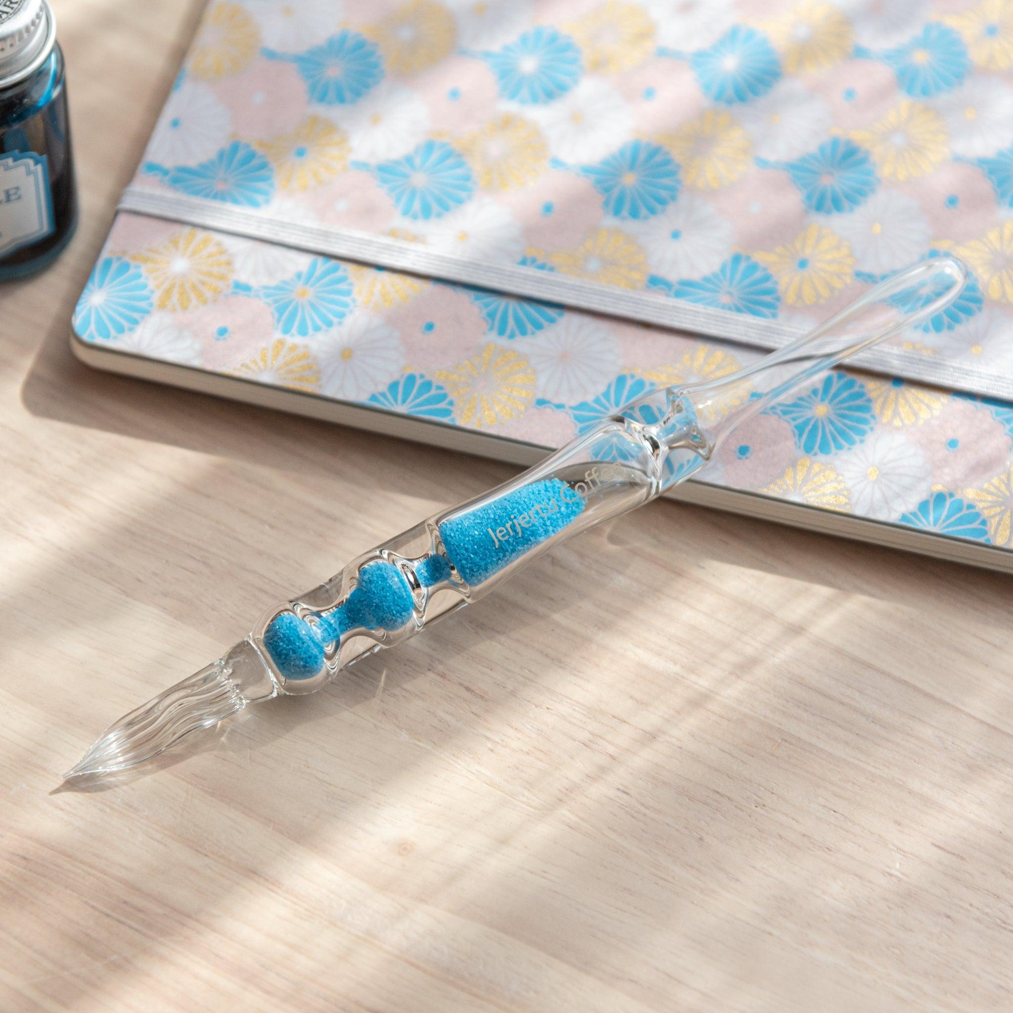 A glass dip pen with blue sand-like particles inside its shaft, engraved with "Jerjertu coffee," rests on a wooden surface near a pastel floral-patterned notebook. A bottle of blue ink is partially visible on the left.