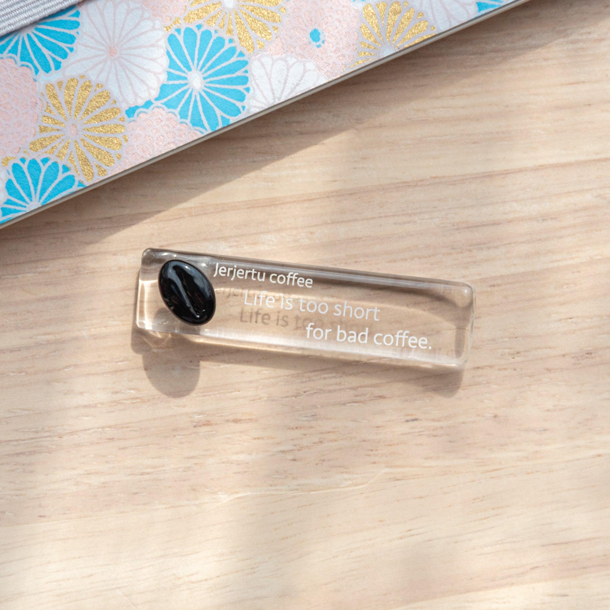 A small glass pen rest inscribed with "Jerjertu coffee, Life is too short for bad coffee" is placed on a wooden surface near a patterned notebook with pastel floral designs.