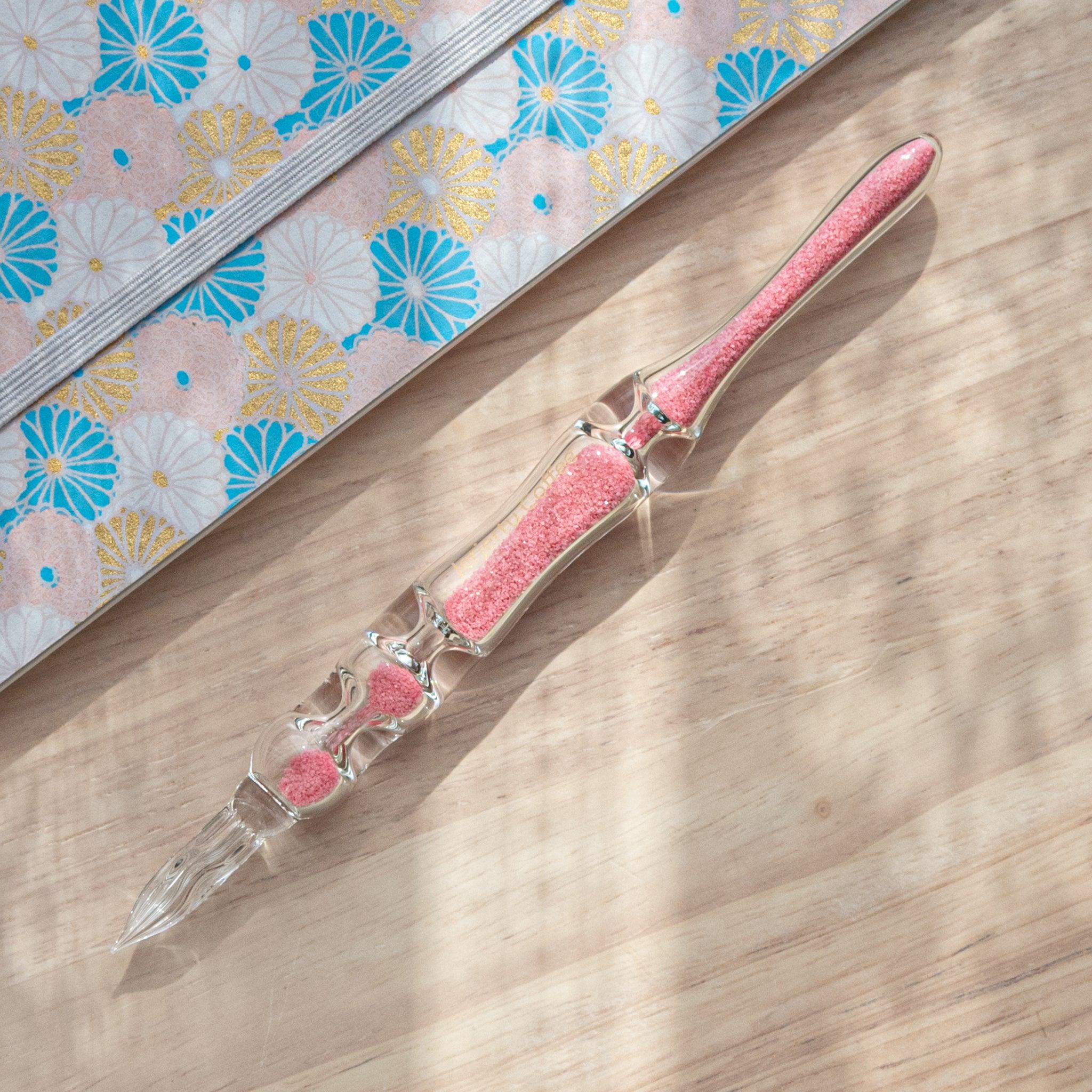 A glass dip pen with pink sand-like particles inside its shaft rests on a wooden surface beside a pastel floral-patterned notebook. The pen's transparent design highlights the vibrant pink particles inside.