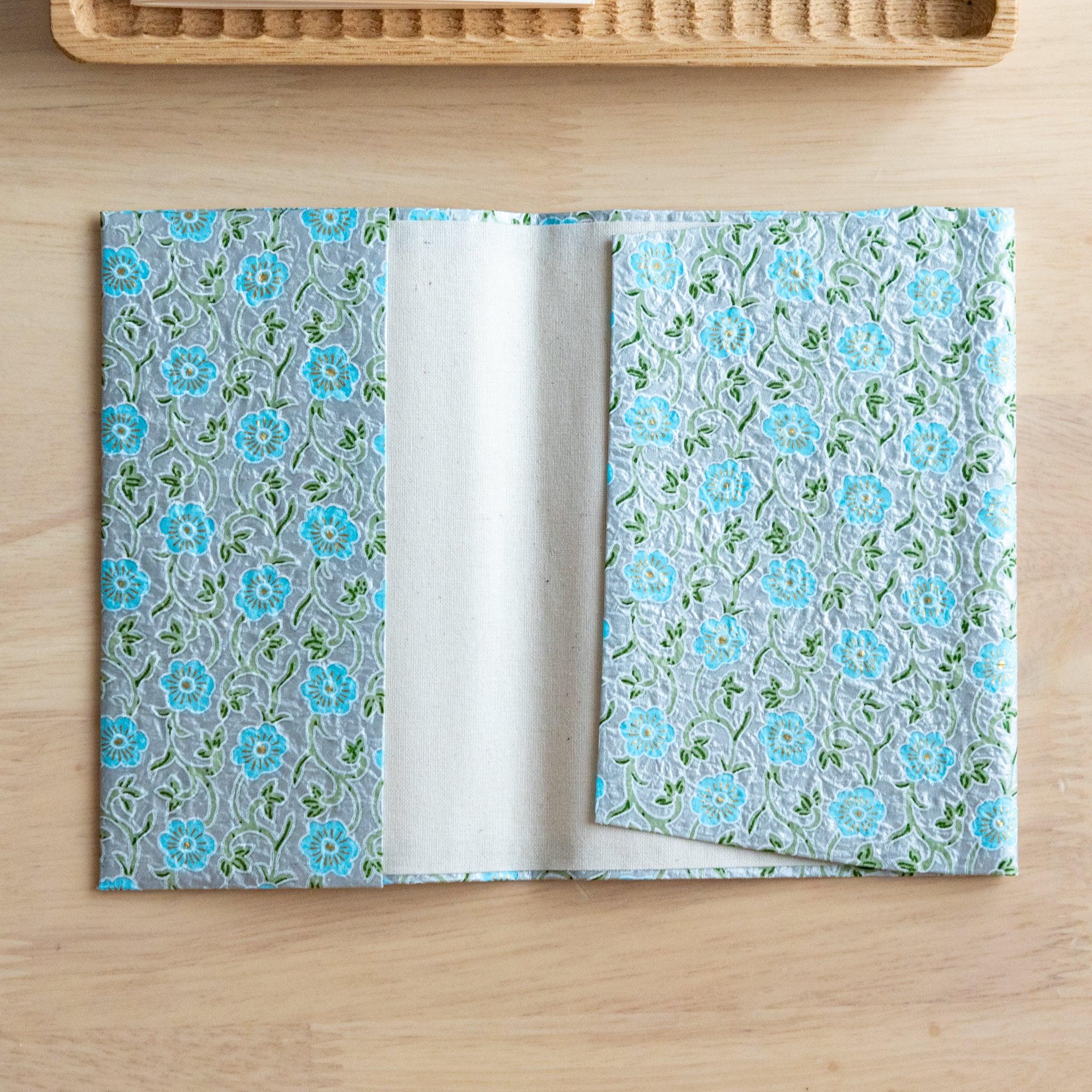 An open notebook cover with a blue floral pattern featuring delicate green vines, revealing its inner white lining on a wooden surface.