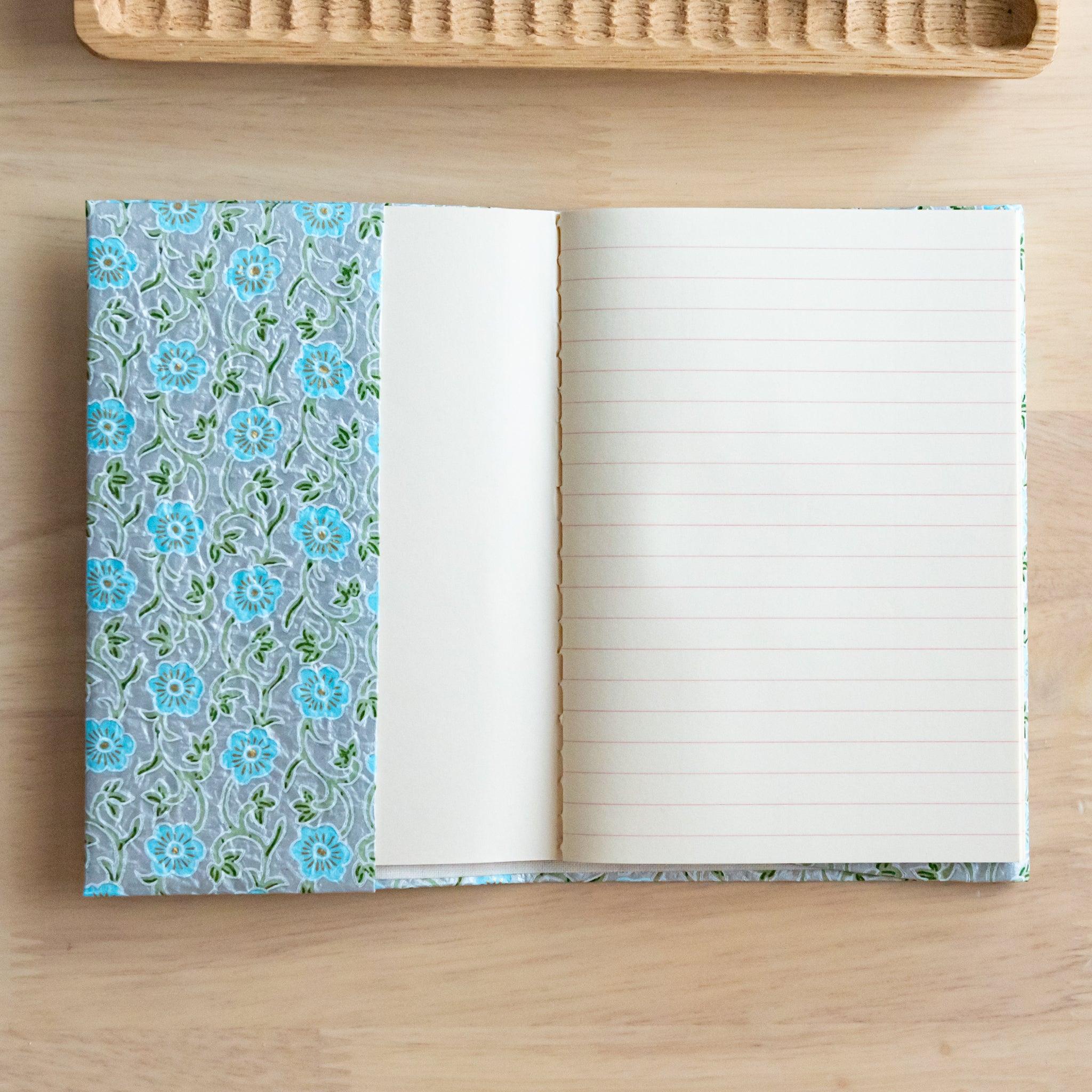 An open notebook with a blue floral patterned cover, showing one lined page and one blank page, placed on a wooden surface.