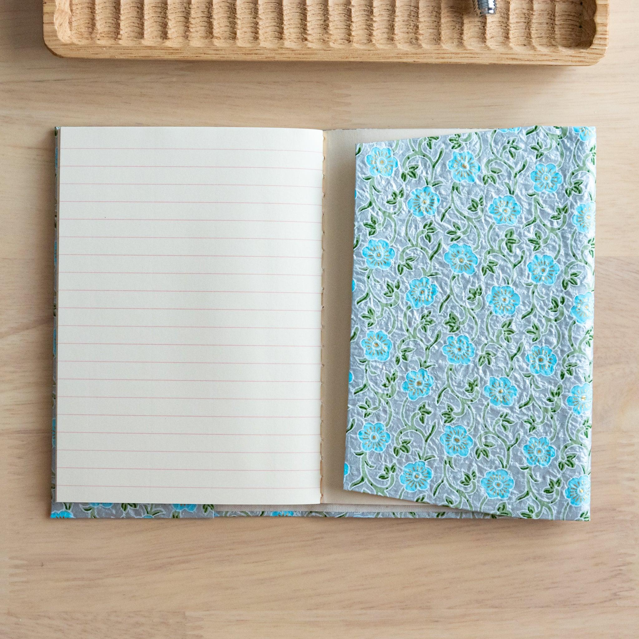 An open notebook with a blue floral patterned cover, revealing a lined page and an inner pocket, placed on a wooden surface.