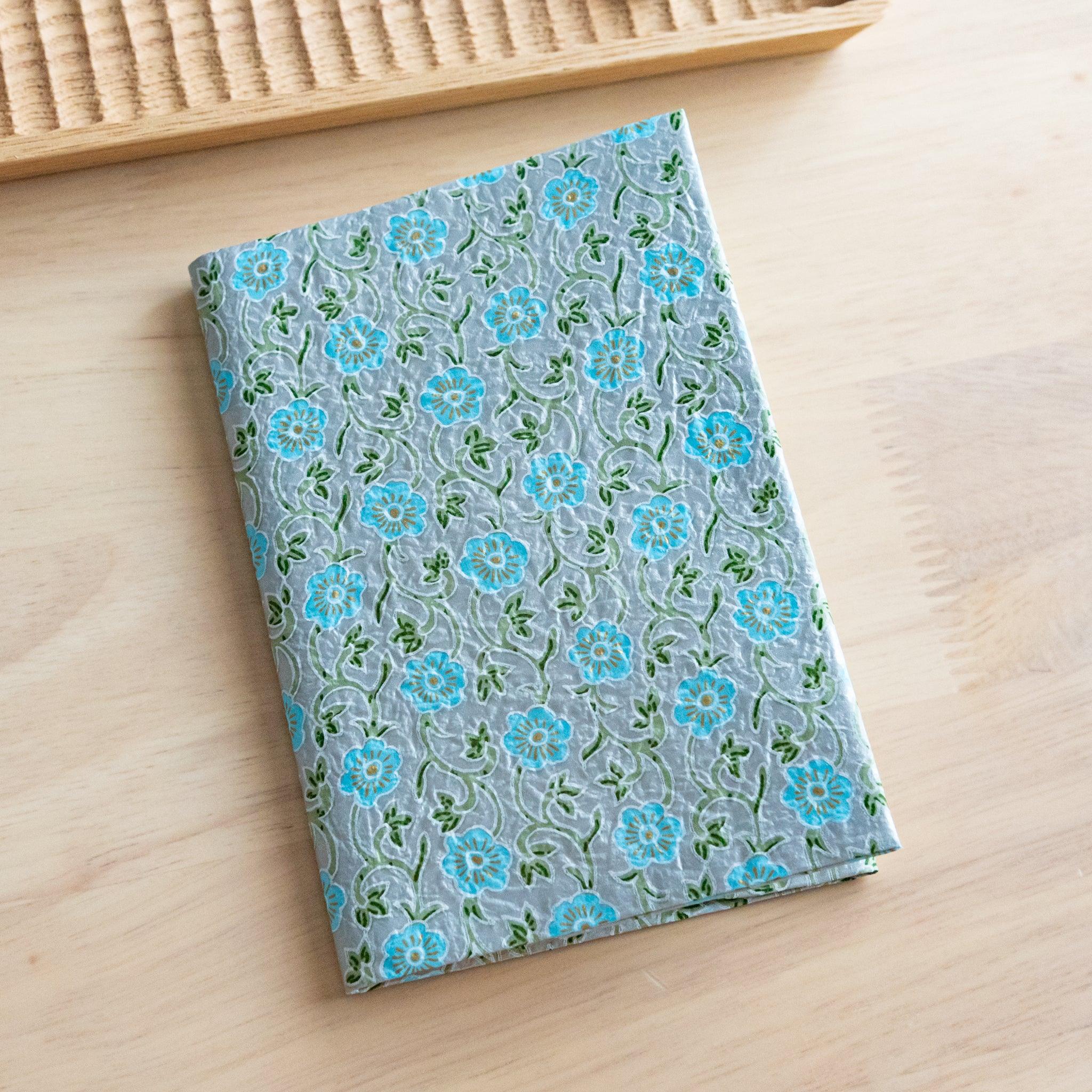 A notebook with a blue floral pattern featuring delicate green vines, placed on a light wooden surface next to a wooden tray.