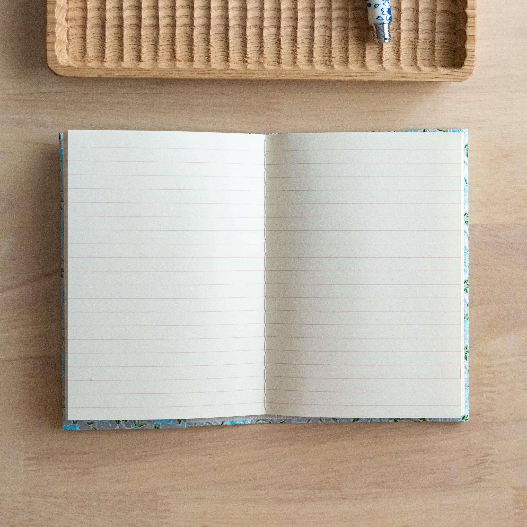 An open notebook with a blue floral patterned cover, fully revealing two lined pages, placed on a light wooden surface.